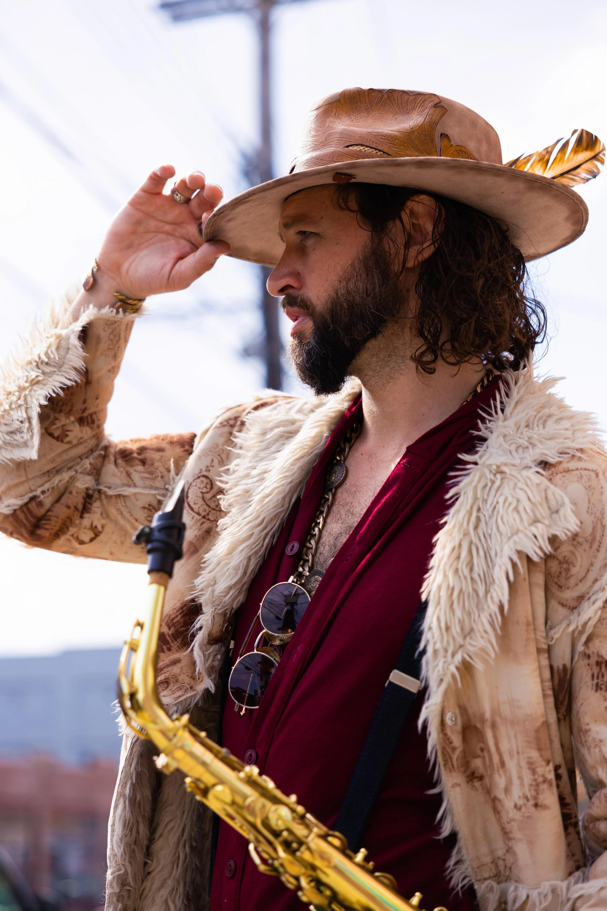 Man with curly hair and beard wearing a large beige hat with feather, sunglasses hanging from red shirt, fur-lined coat, holding a saxophone, outdoors.