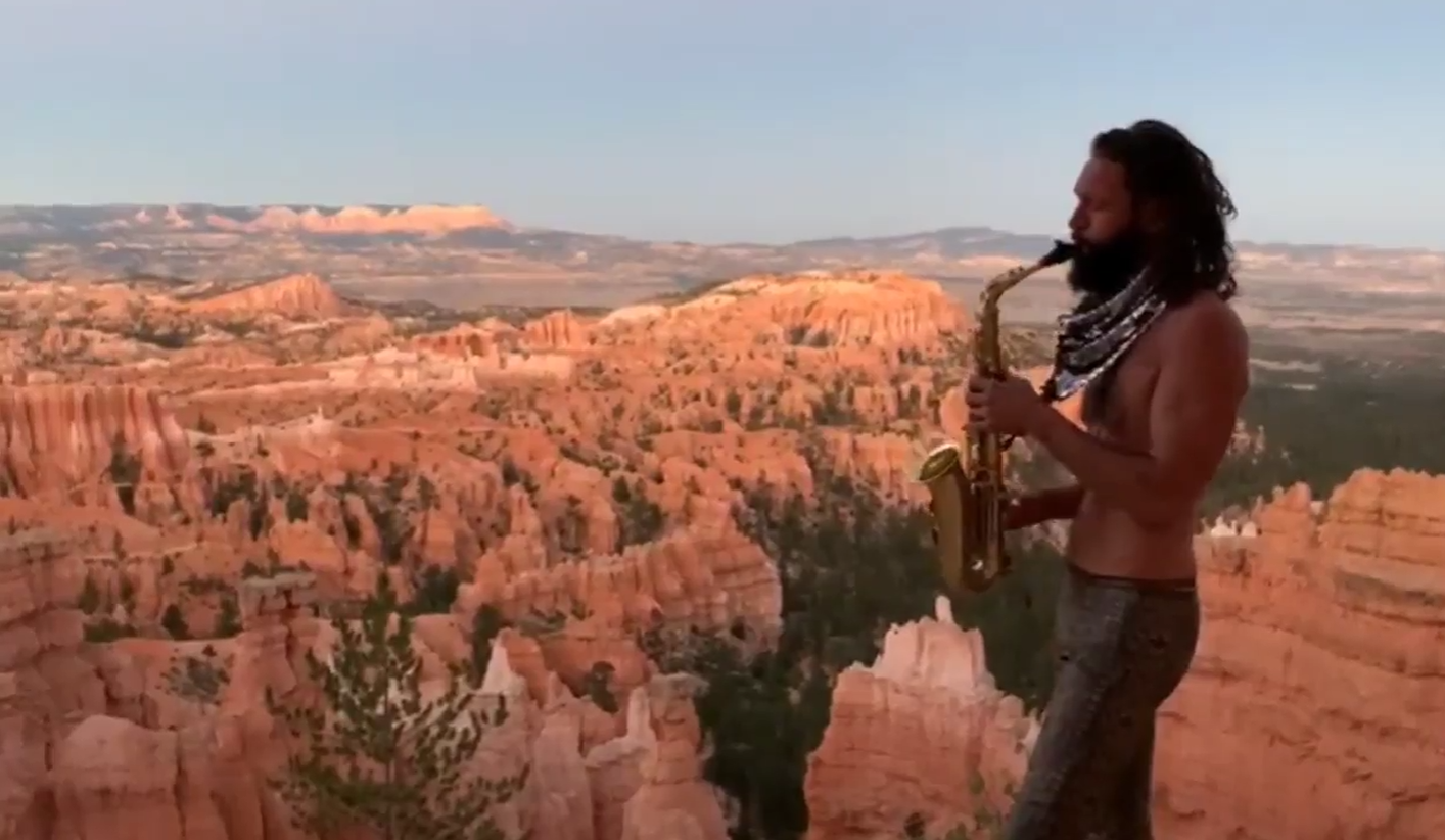 A man with long hair and a beard playing a saxophone on a ledge overlooking the Grand Canyon during sunset.