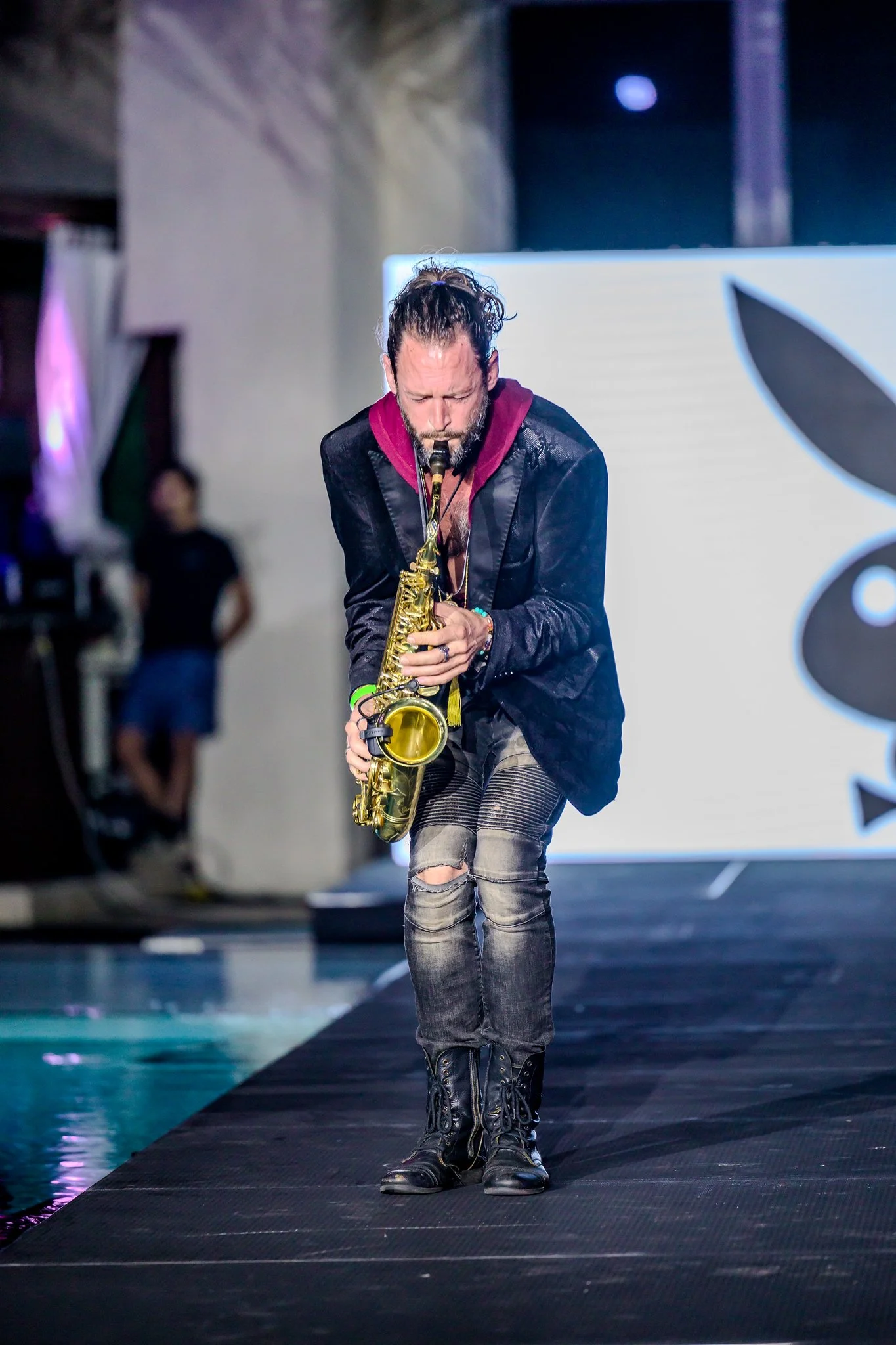 A man with long hair and a beard playing a gold saxophone on a stage with a large white backdrop featuring a black butterfly logo.