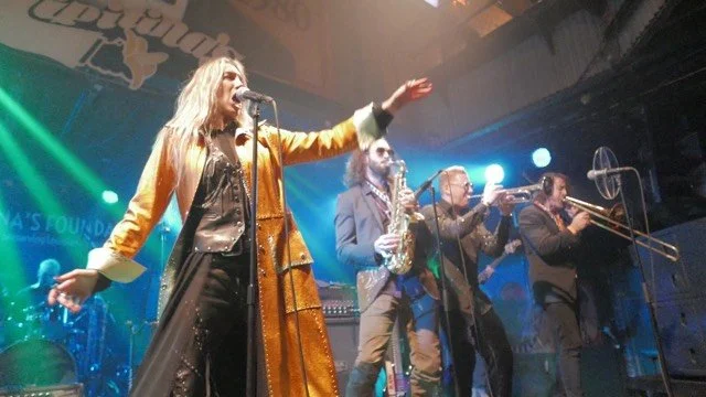 A band performs on stage with a female singer in the foreground, wearing a gold coat and black outfit, and three male musicians playing saxophone, trumpet, and trombone in the background.