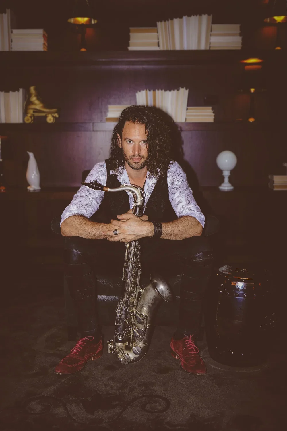 A man with long, curly dark hair and a serious expression is sitting on a black chair, holding a saxophone. He is wearing a white patterned shirt, a black vest, black pants with knee patches, and red shoes. The background features dark wood shelves w