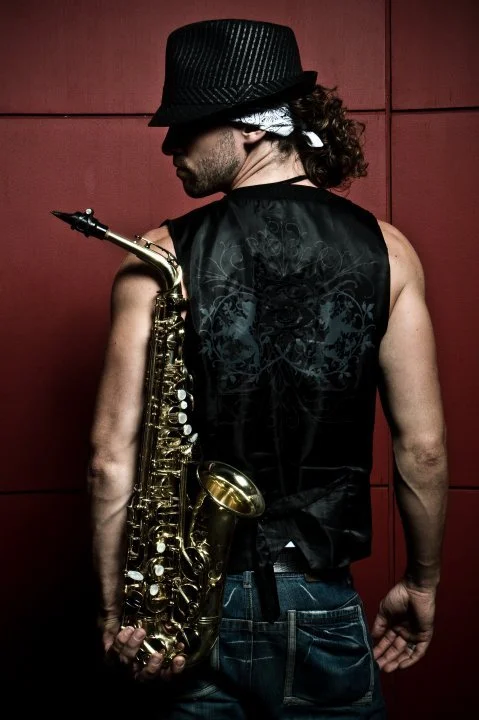 A man with long curly hair wearing a black fedora hat, sleeveless black shirt with floral pattern, and denim jeans, holding a saxophone on his back against a red wall.