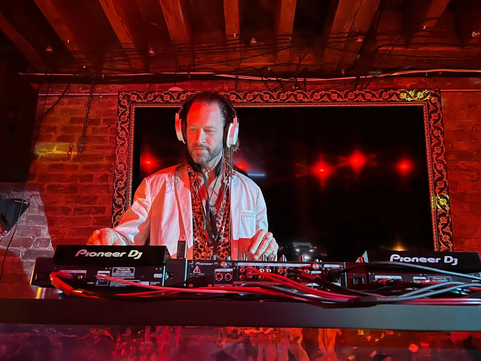 A DJ wearing headphones and a white jacket, with a leopard print shirt underneath, is performing at a nightclub with red lighting and a brick wall in the background.