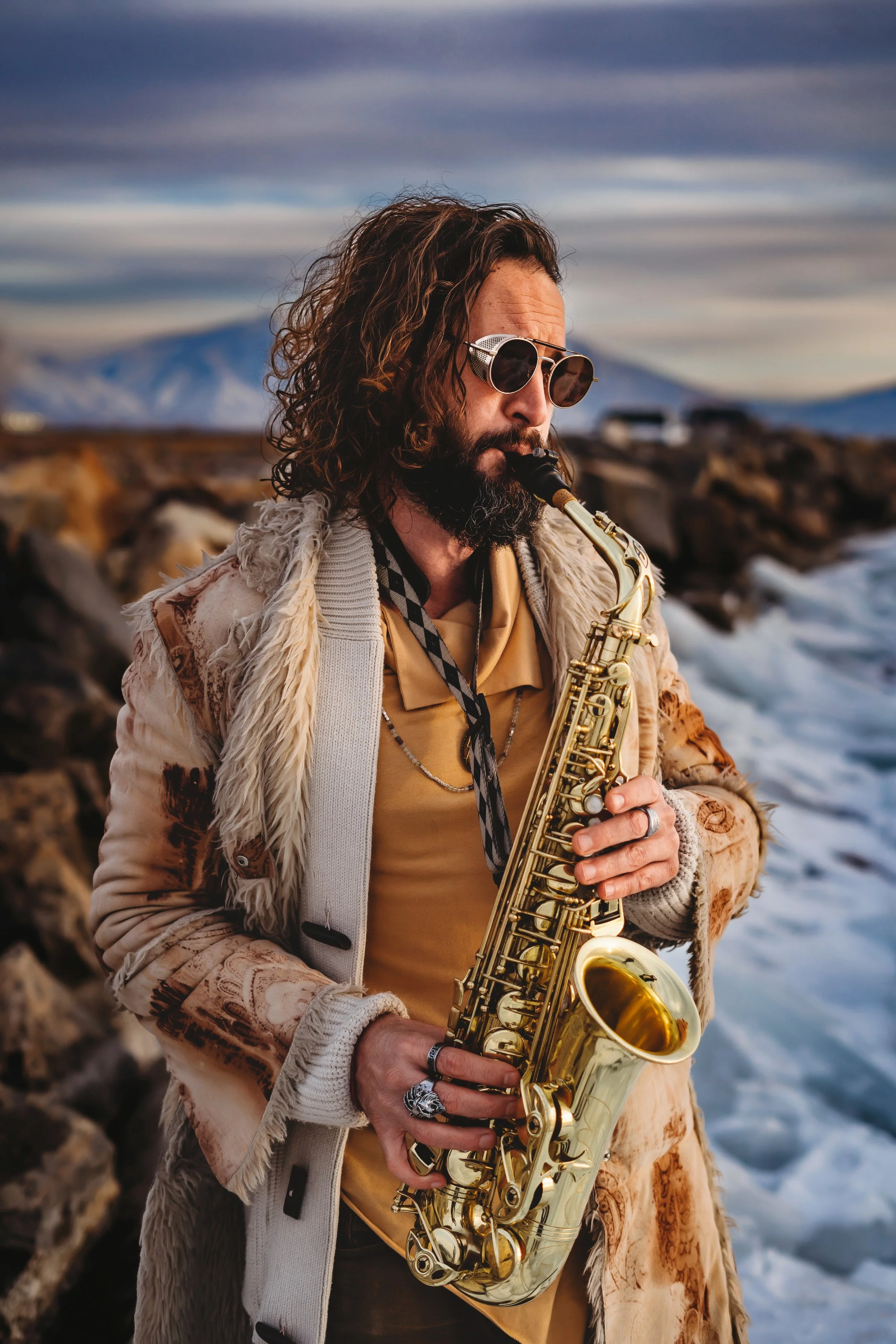 A man with curly hair and sunglasses playing a saxophone outdoors near the ocean with mountains in the background.