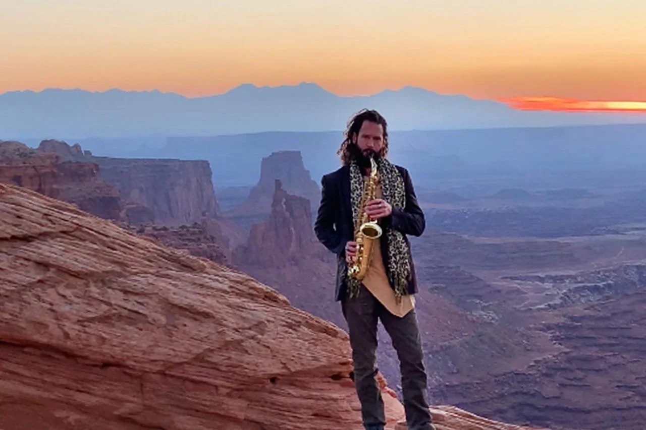 A man with long hair and a beard playing a saxophone on a rocky ledge at sunset, with the Grand Canyon in the background.