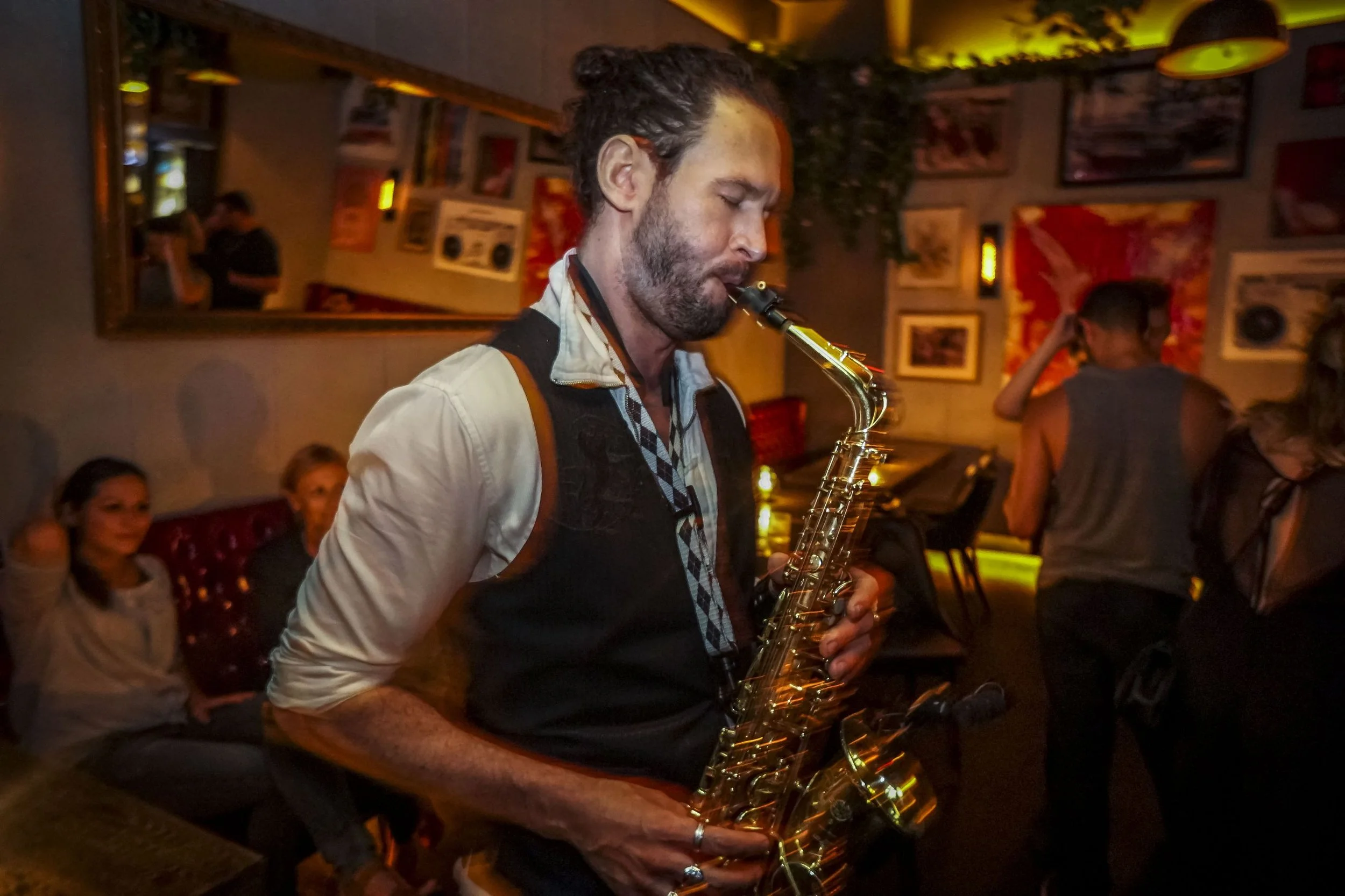 A man with closed eyes playing a saxophone in a dimly lit bar or restaurant, surrounded by people and decorated with various artwork.