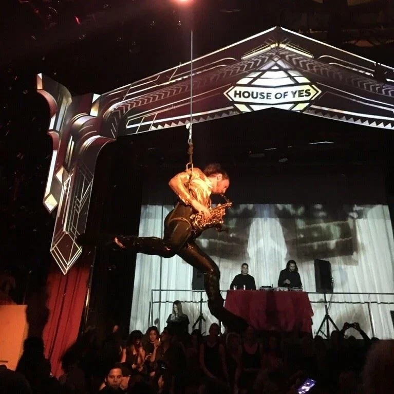 A performer hanging on a harness and playing a saxophone on stage at House of Yes, with a DJ setup in the background and audience watching.