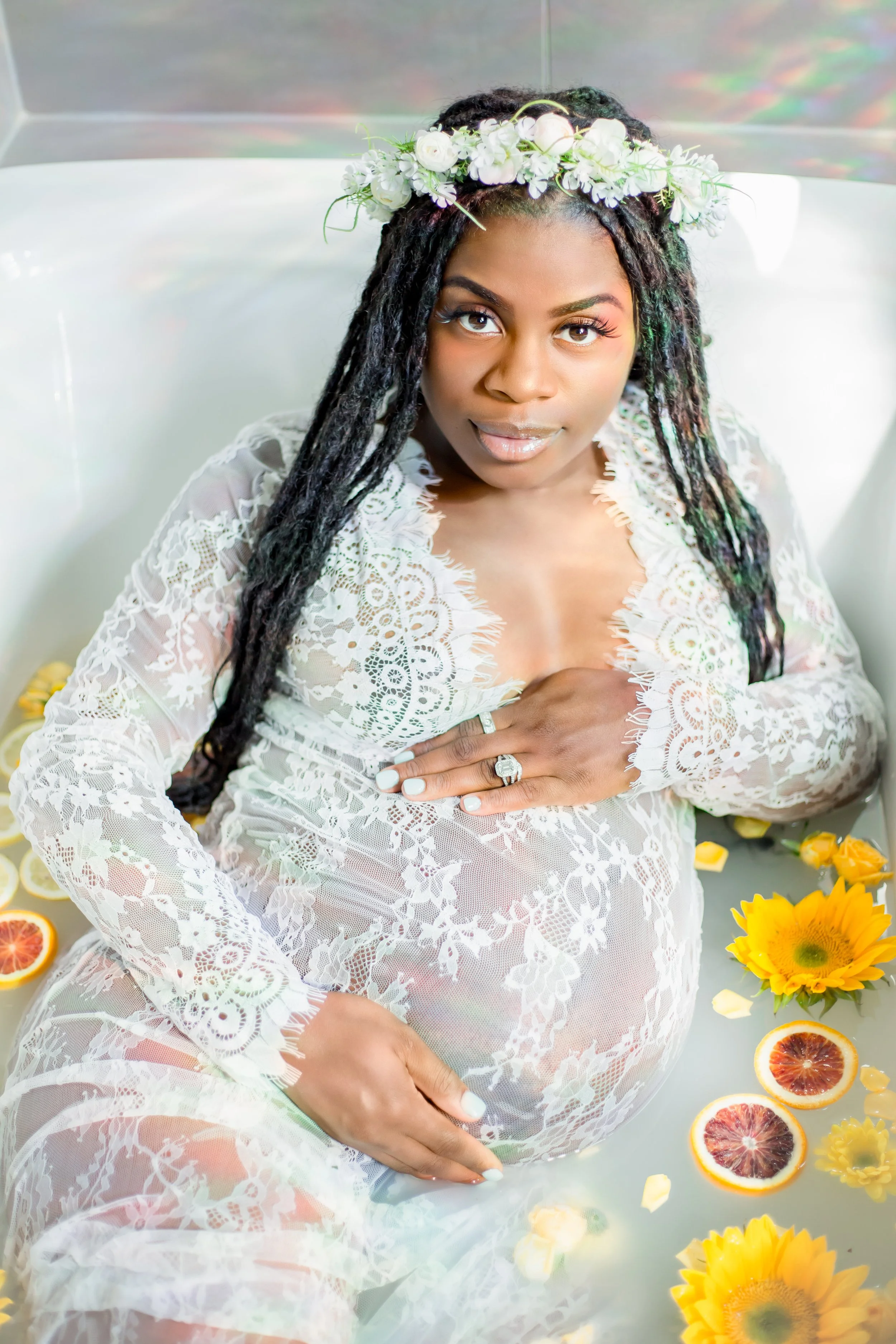 A pregnant woman with long dreadlocks and a flower crown, wearing a white lace dress, lies in a bathtub filled with water, flower petals, and sliced citrus fruits, looking at the camera.