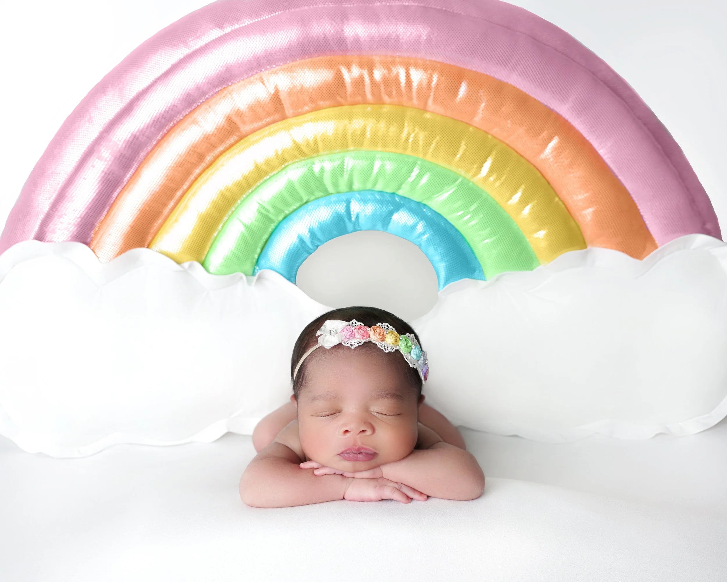 A sleeping baby with a floral headband resting on a white surface, positioned in front of a colorful rainbow backdrop with clouds.
