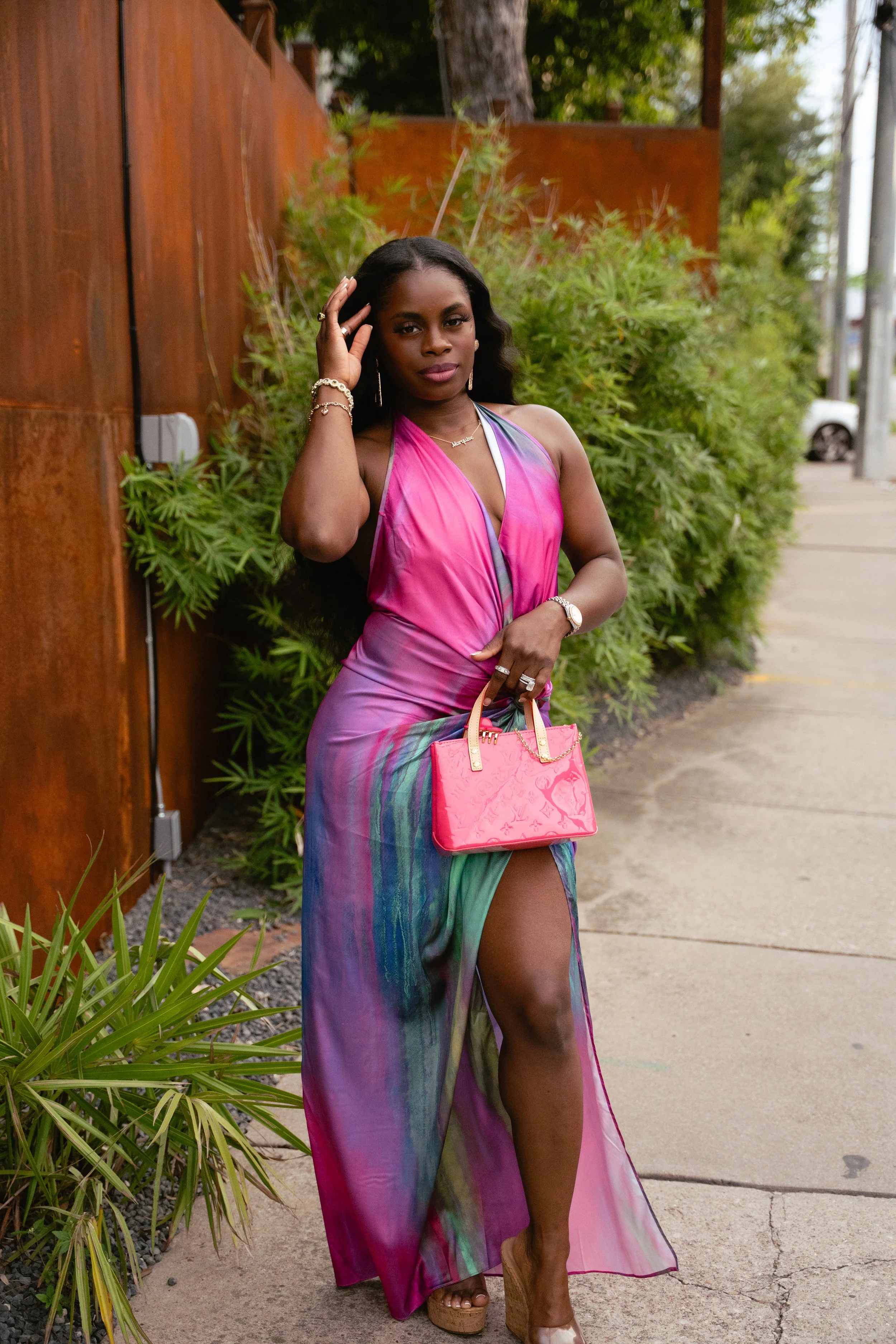 A woman stands outdoors on a sidewalk with lush greenery and a rusty fence in the background. She wears a colorful, pink and green flowing dress with a slit, beige platform heels, earrings, and bracelets. She holds a pink handbag and has long black hair.