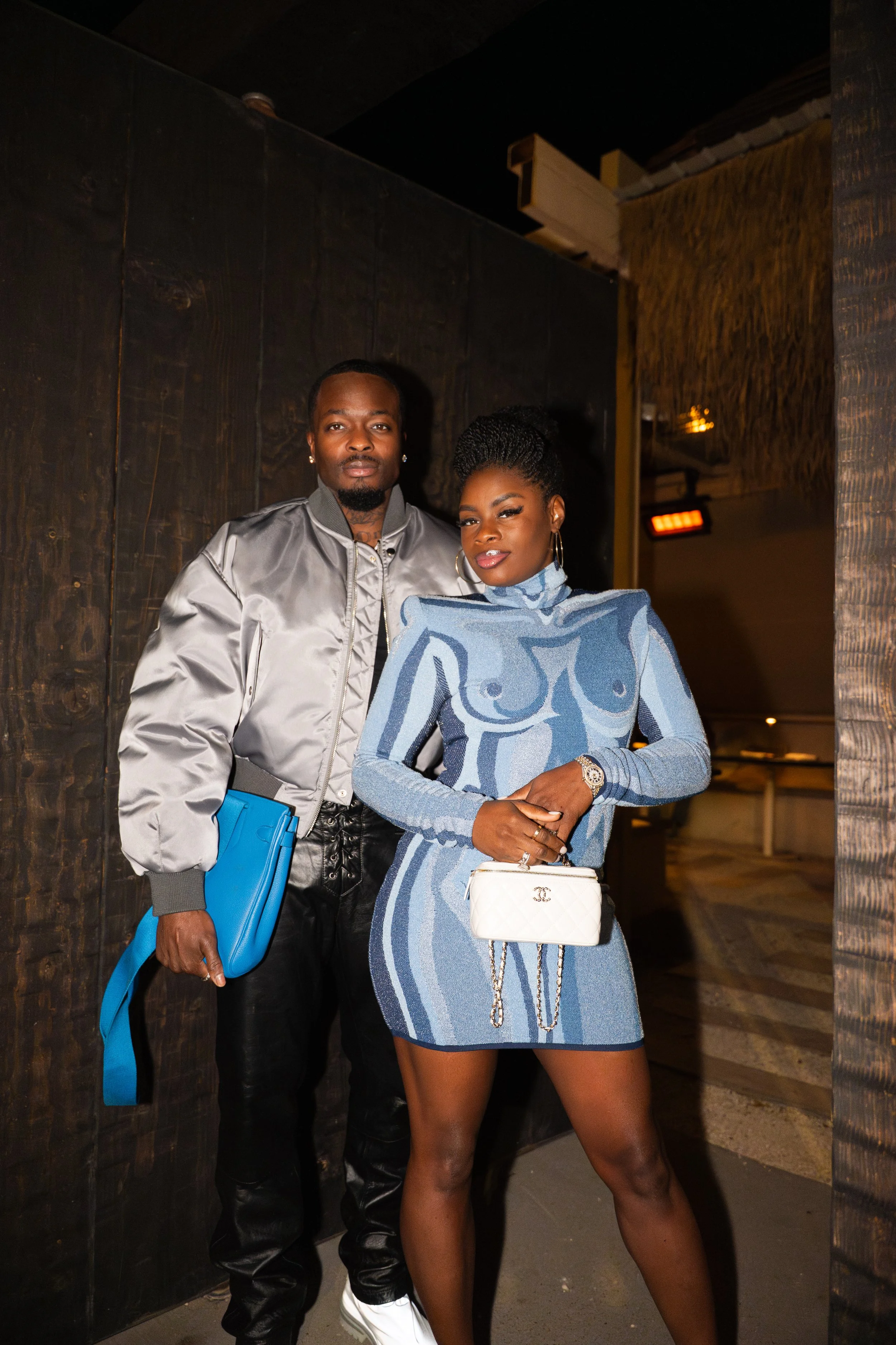 A man and woman standing together indoors, dressed stylishly, with the woman holding a small white handbag, both posing for the photo.