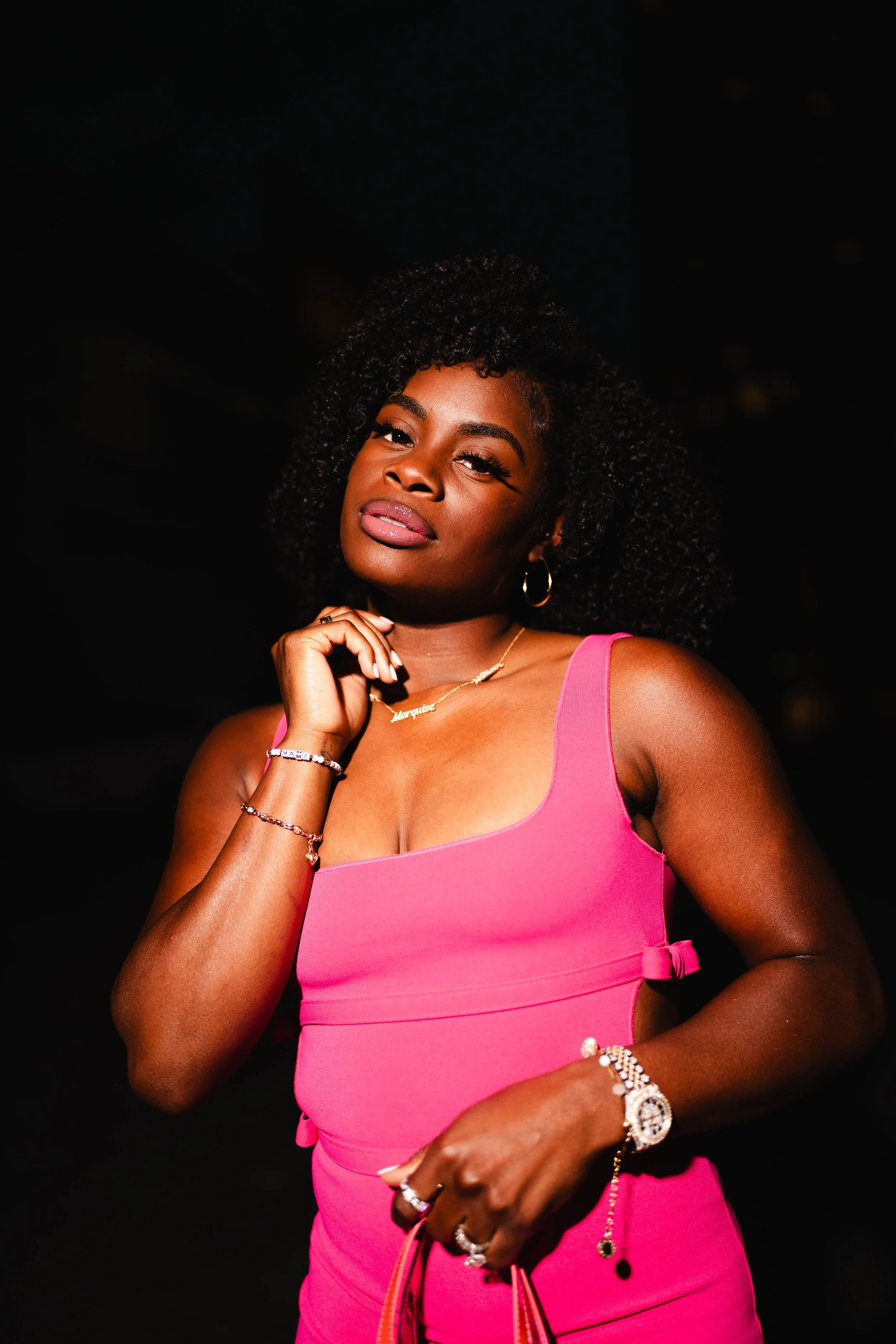 A young Black woman with curly hair in a pink dress, wearing jewelry and posing confidently against a dark background.