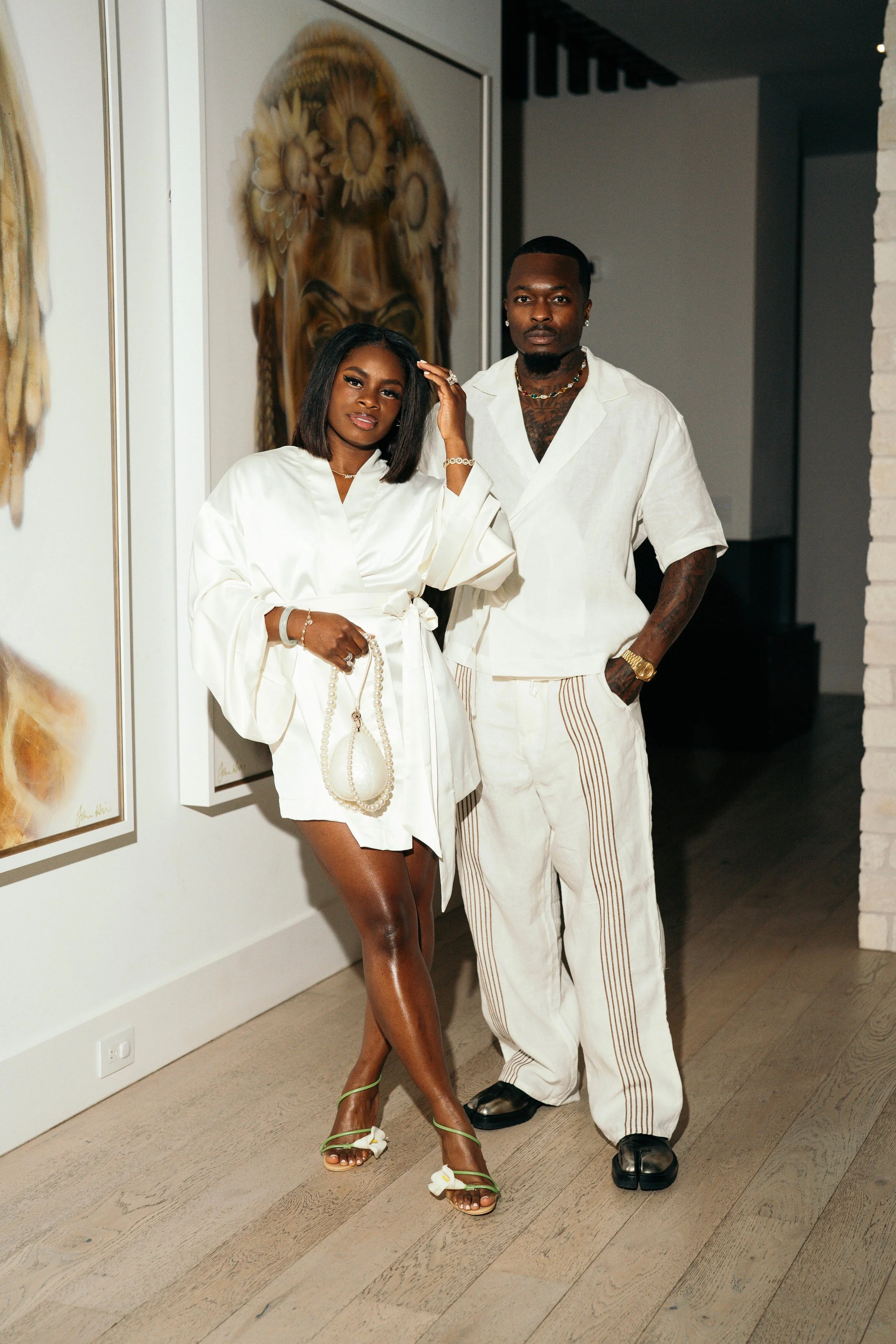 A woman and a man posing together indoors. The woman is wearing a white satin robe, holding a small pearl handbag, and wearing green heels with bows. The man is dressed in a loose white shirt and white pants with side stripes. Both have jewelry, and the woman has her hair styled in a bob. Behind them are two artworks depicted in a gallery setting.