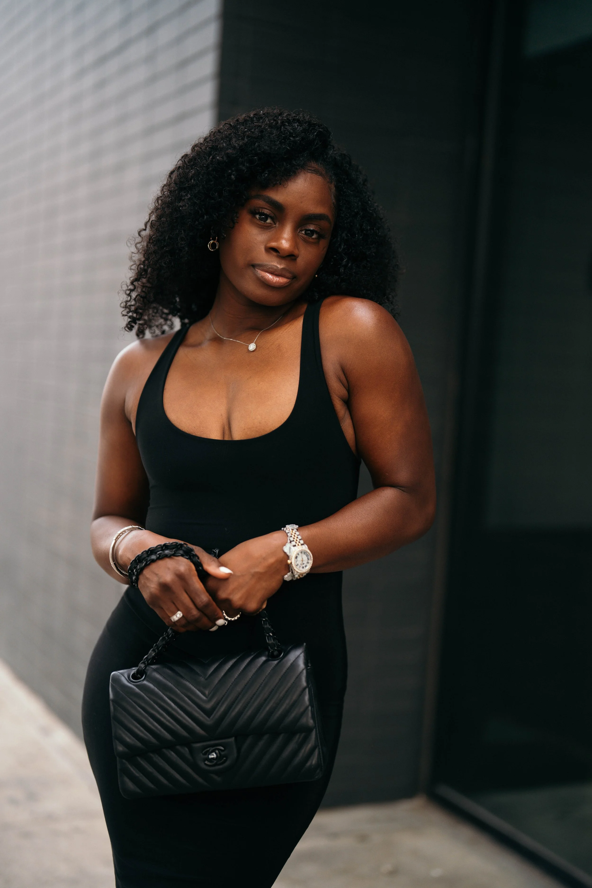 A woman with dark skin and curly hair wearing a black sleeveless dress, jewelry, and holding a black Chanel purse, standing against a dark wall.