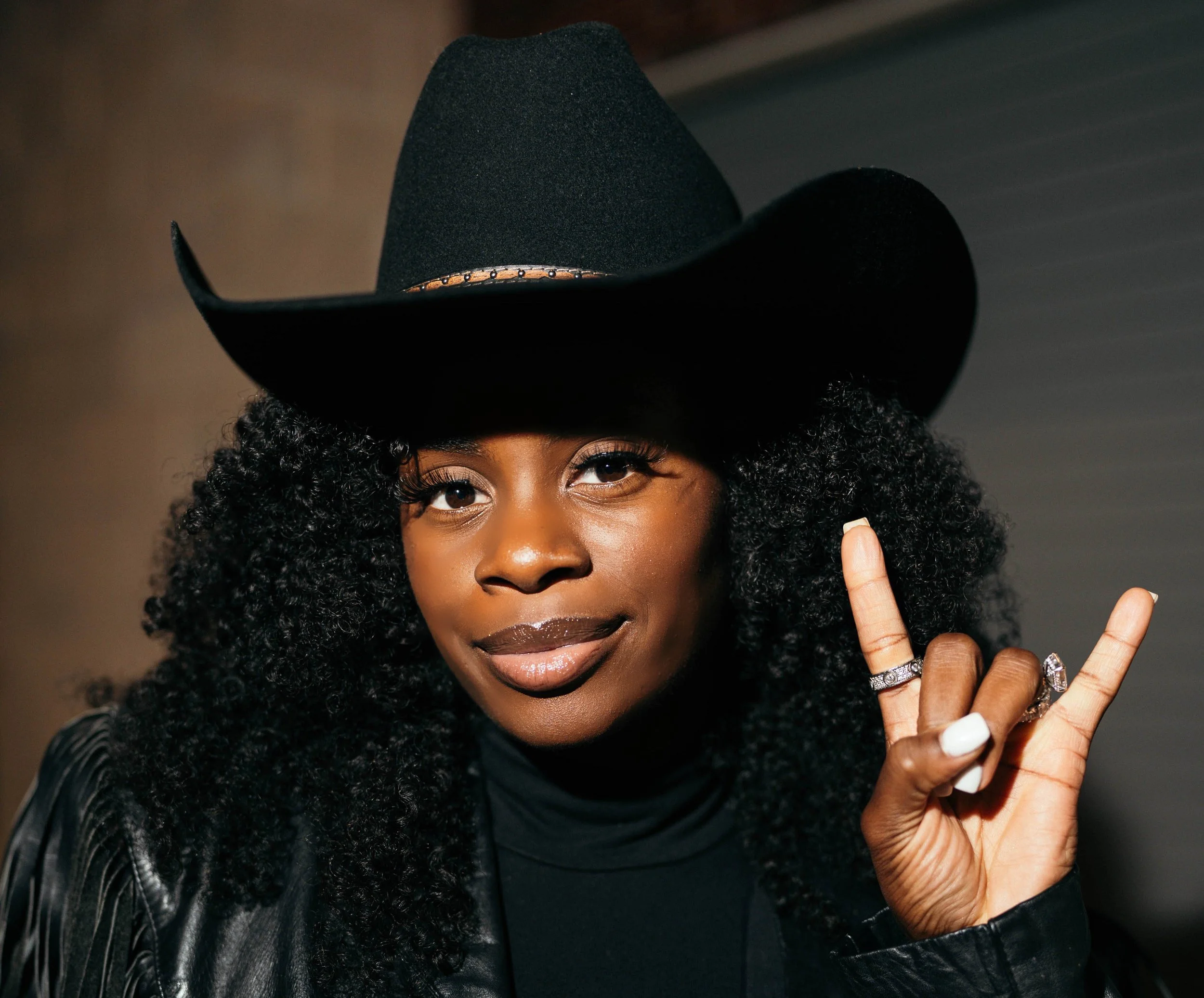 A woman with dark curly hair wearing a black cowboy hat, black turtleneck, and black leather jacket, making a 'rock on' gesture with her right hand.
