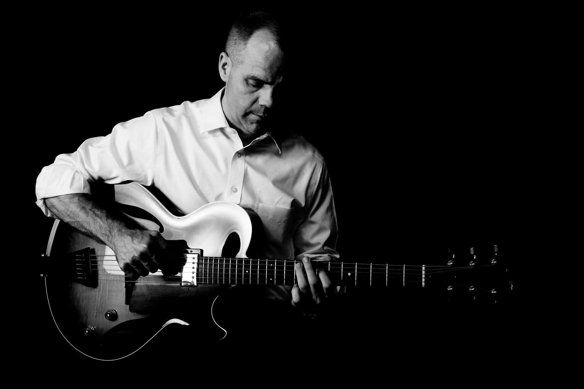 A black and white photo of a man in a white shirt playing an electric guitar against a dark background.