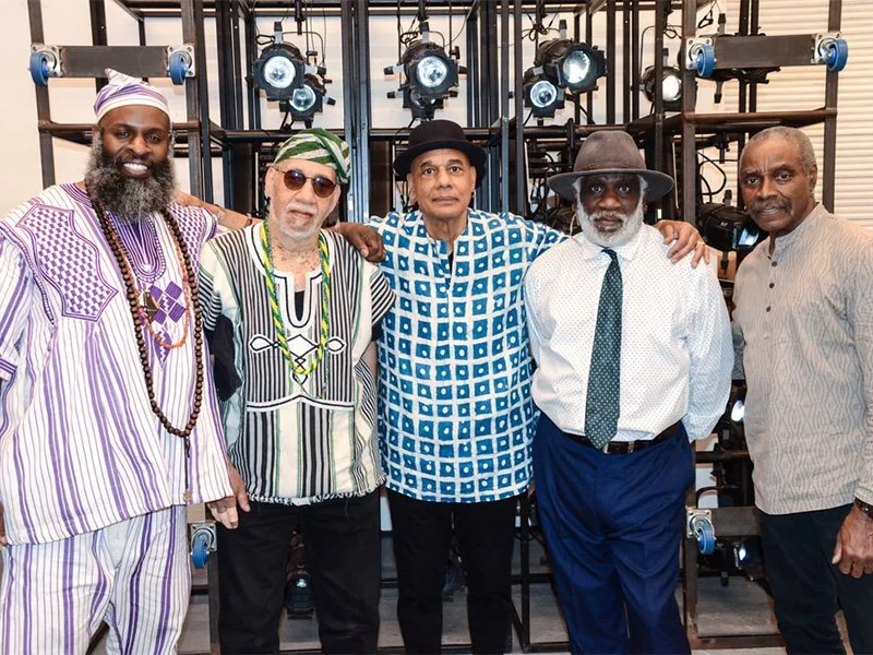 Five men standing together in front of theatrical stage lights, dressed in colorful and patterned clothing, posing for a photo.