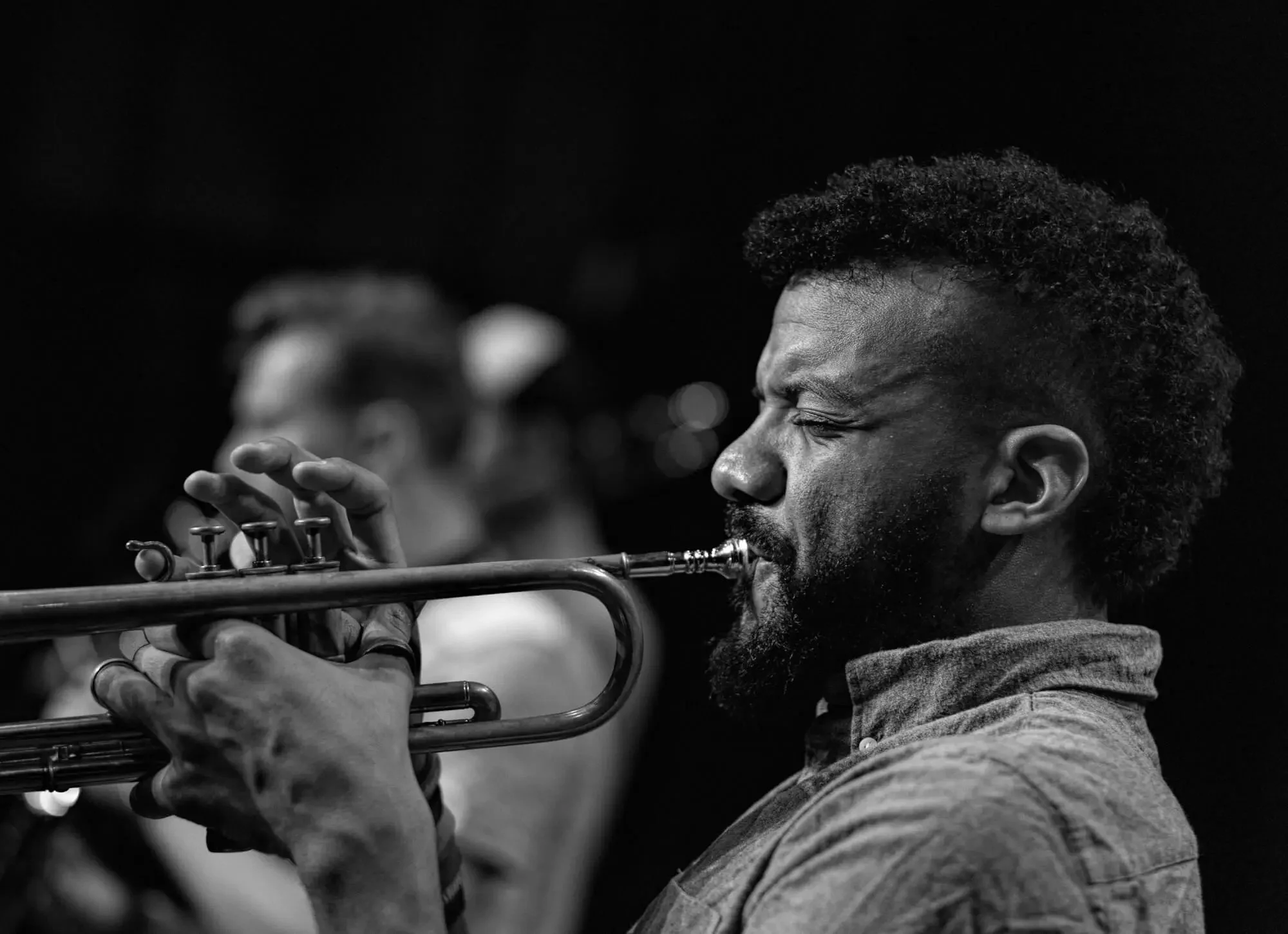 A black and white photo of a man playing a trumpet, with his eyes closed and an expression of concentration, seen from the side.