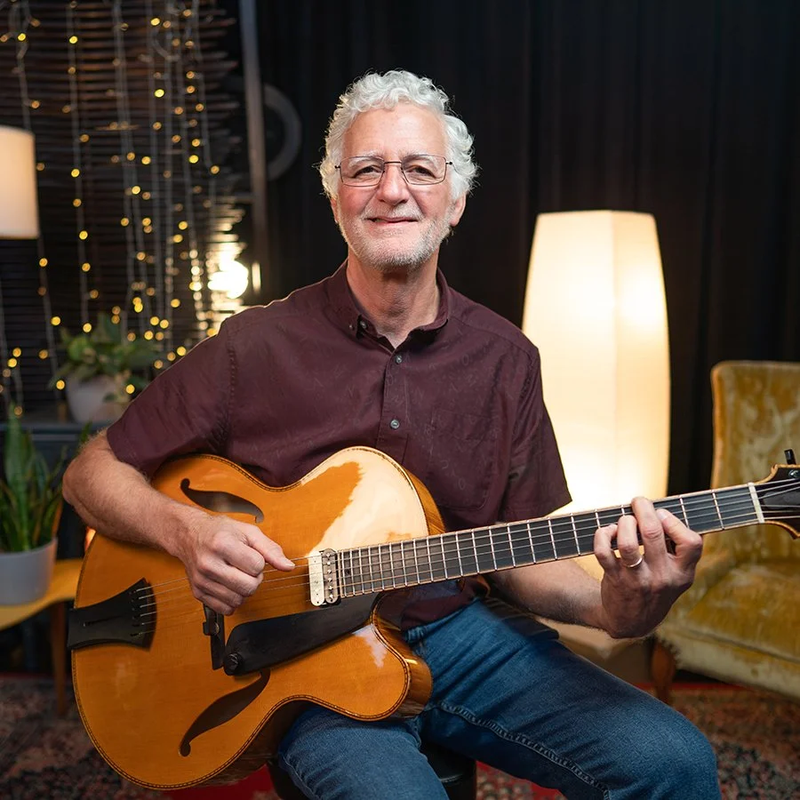 A man with white hair and glasses playing an acoustic guitar indoors, with warm lighting and decorative plants in the background.