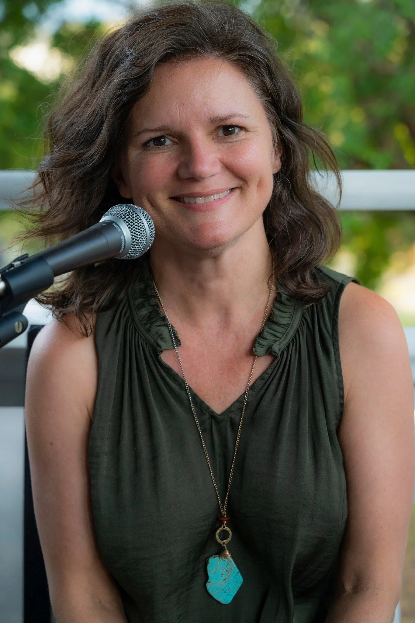 A smiling woman sitting outdoors near a microphone, wearing a dark sleeveless top and a necklace with a large turquoise pendant.