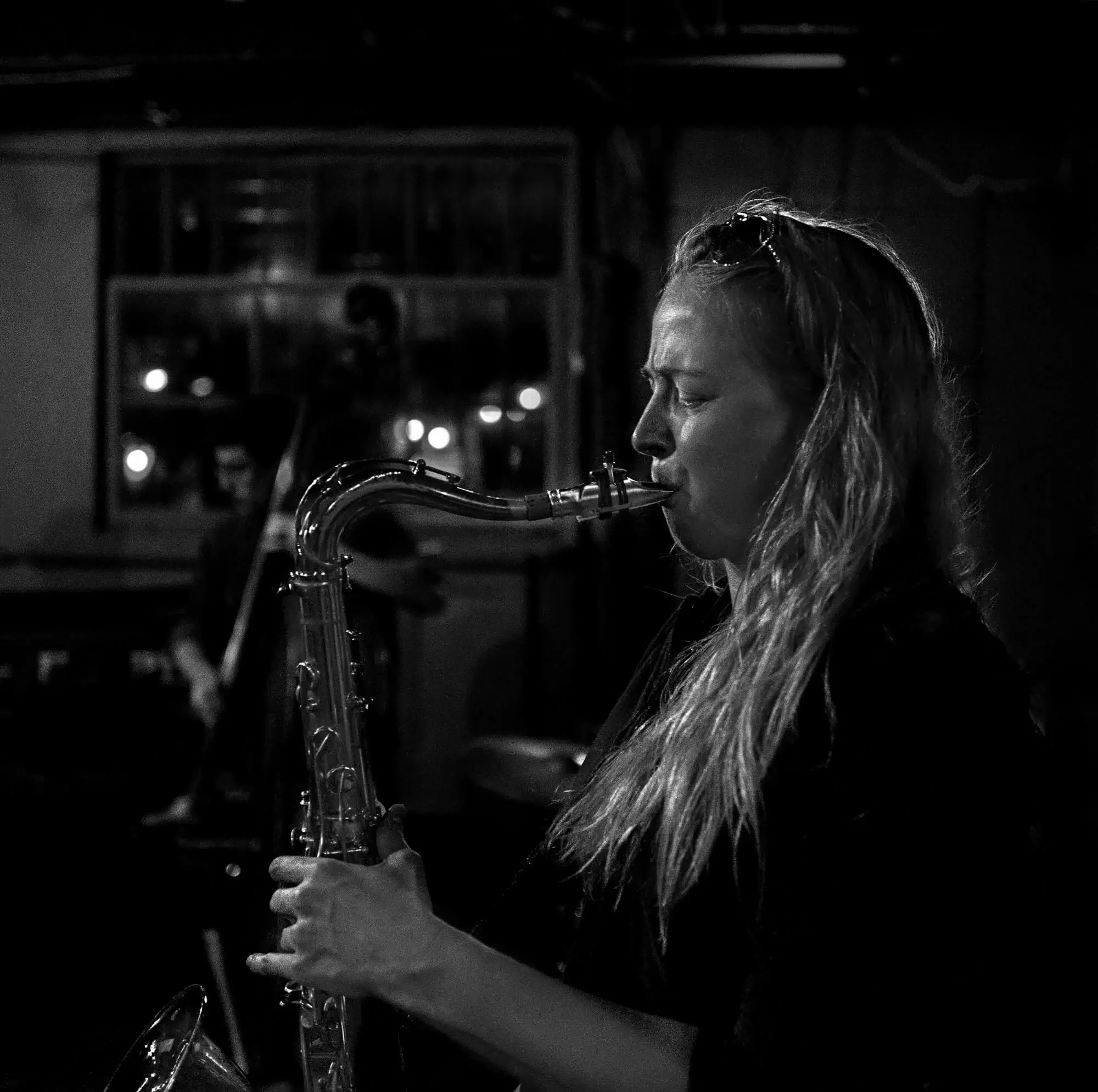 A woman playing the saxophone in a dimly lit room, captured in black and white.