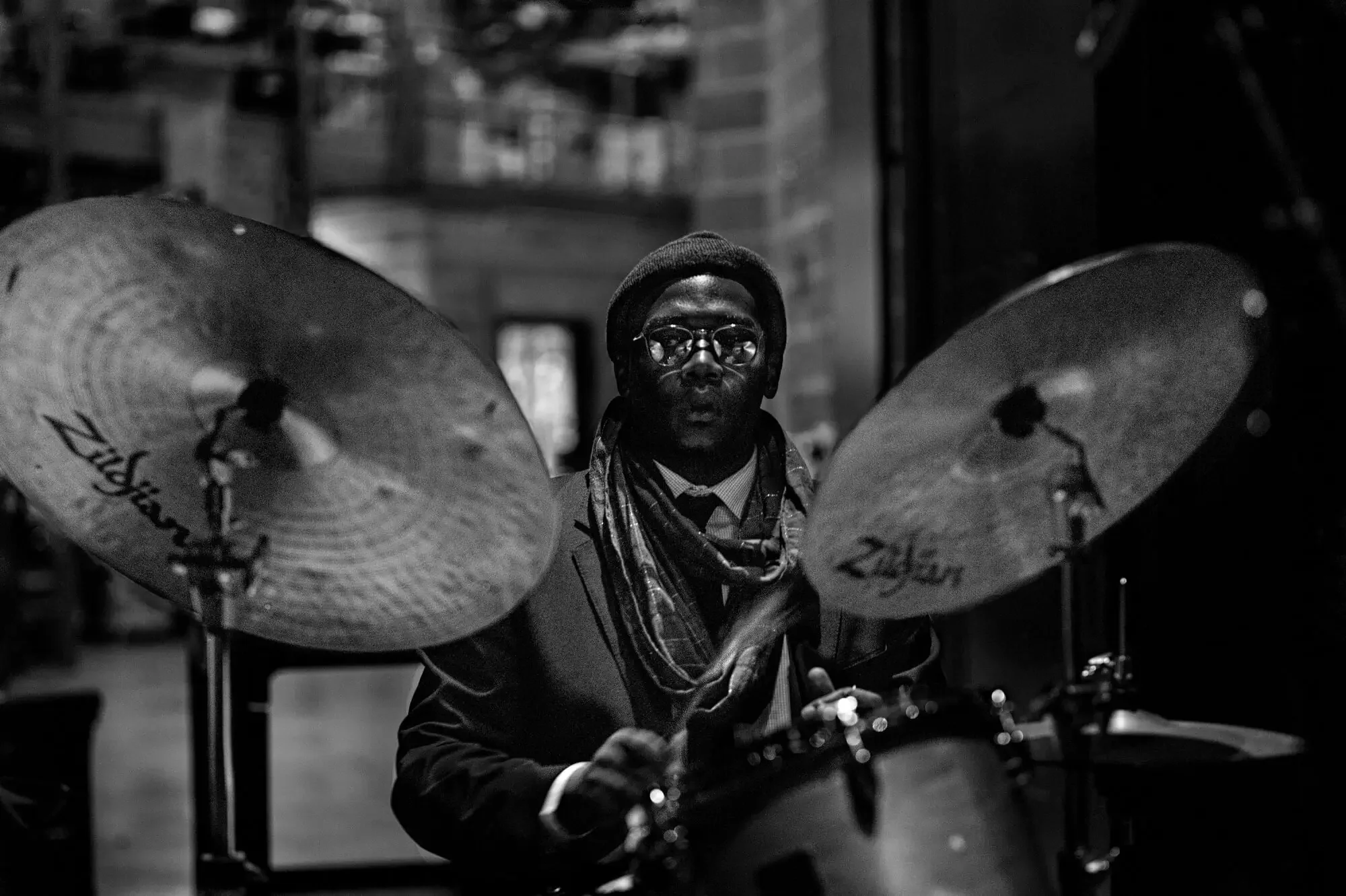 A man playing the drums in a dimly lit indoor space, with a serious expression, wearing glasses, a hat, and a scarf.