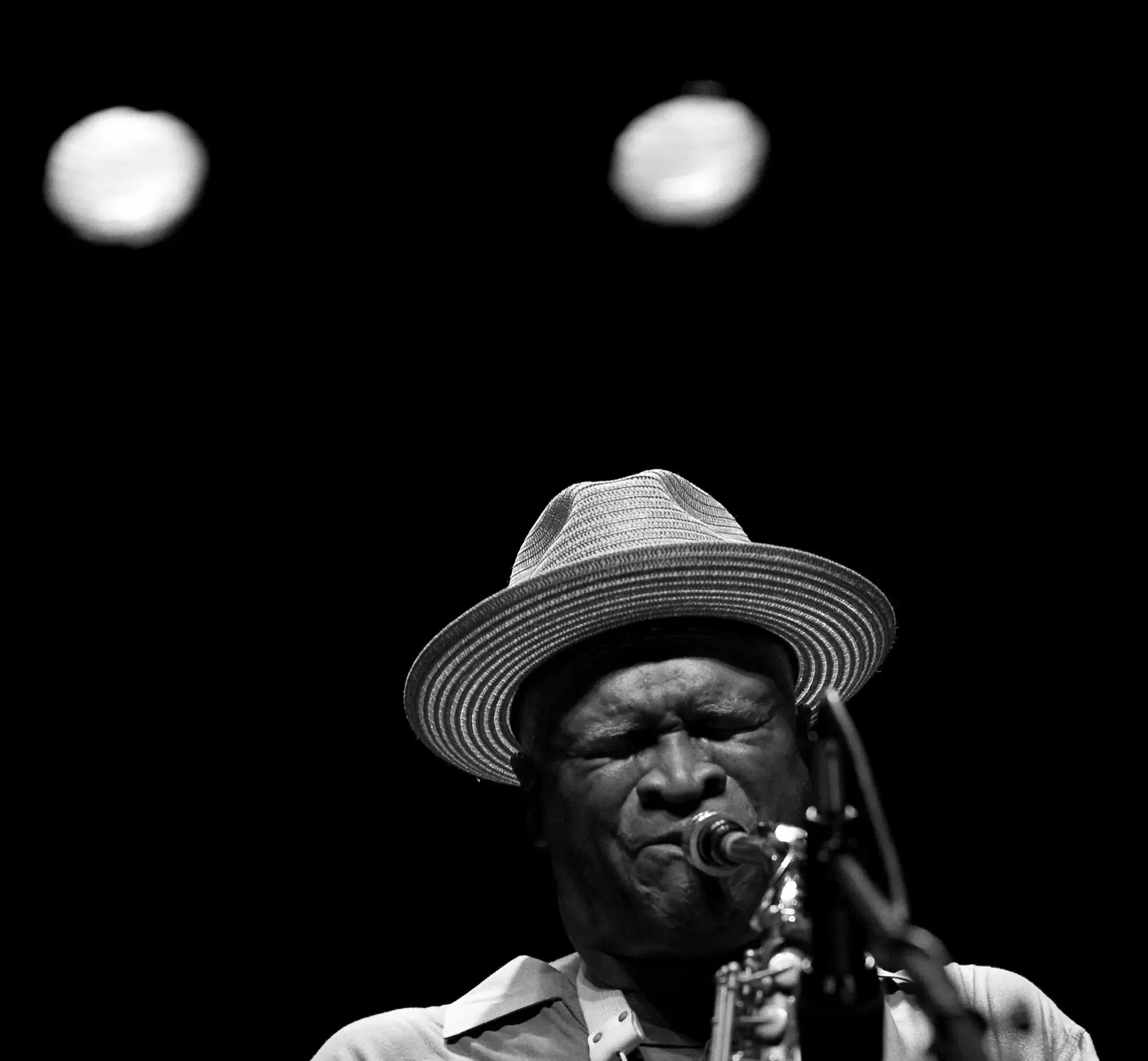 A black-and-white photo of a man wearing a wide-brimmed hat, singing into a microphone with a focused expression.