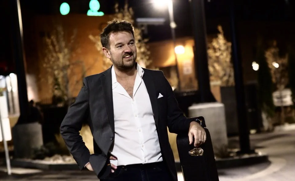 A man in a black suit with a white shirt, holding a guitar case, standing on a street at night with streetlights and trees in the background.