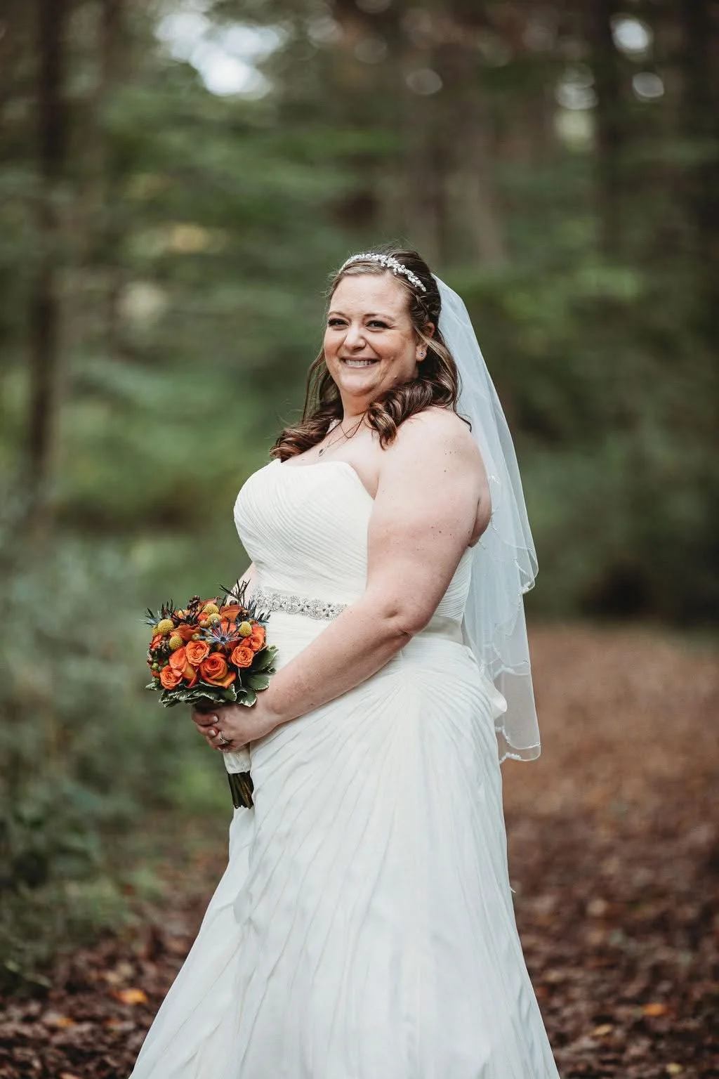 A bride in a white strapless wedding gown holding a bouquet of orange and red flowers, standing outdoors in a wooded area.