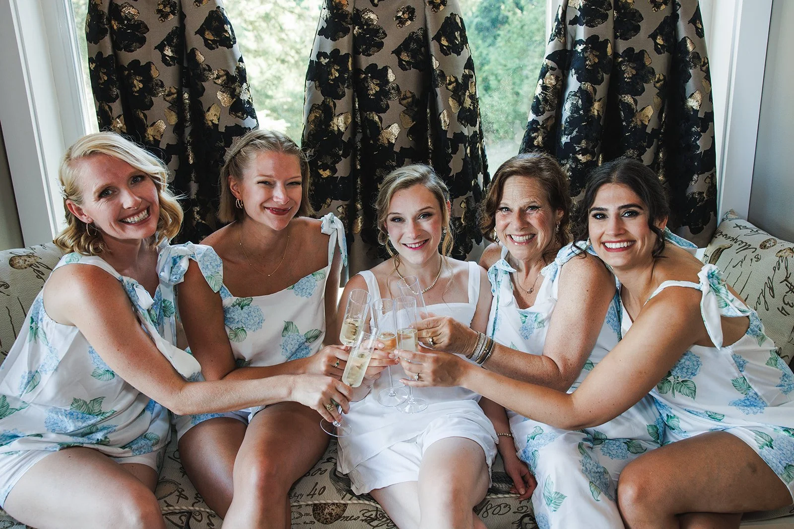 Six women in matching white dresses with blue floral patterns sitting on a couch, holding champagne glasses, toasting and smiling. They are in a well-lit room with dark curtains and greenery outside the window.