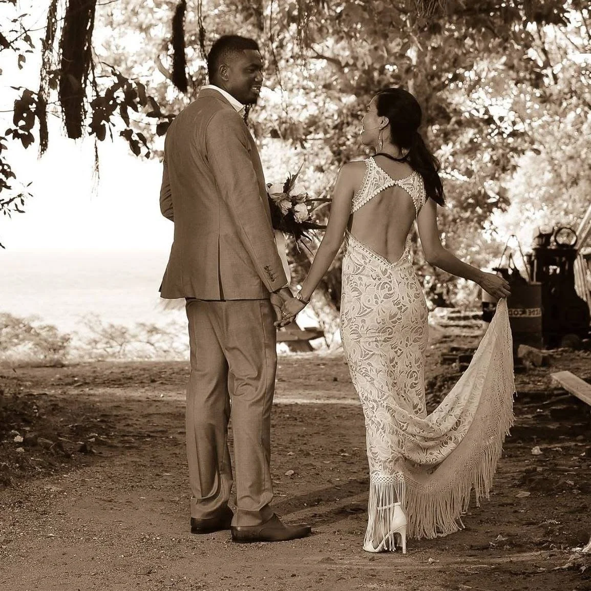 A couple holding hands during their wedding ceremony outdoors, with the man in a suit and the woman in a lace dress holding a bouquet, standing under trees.