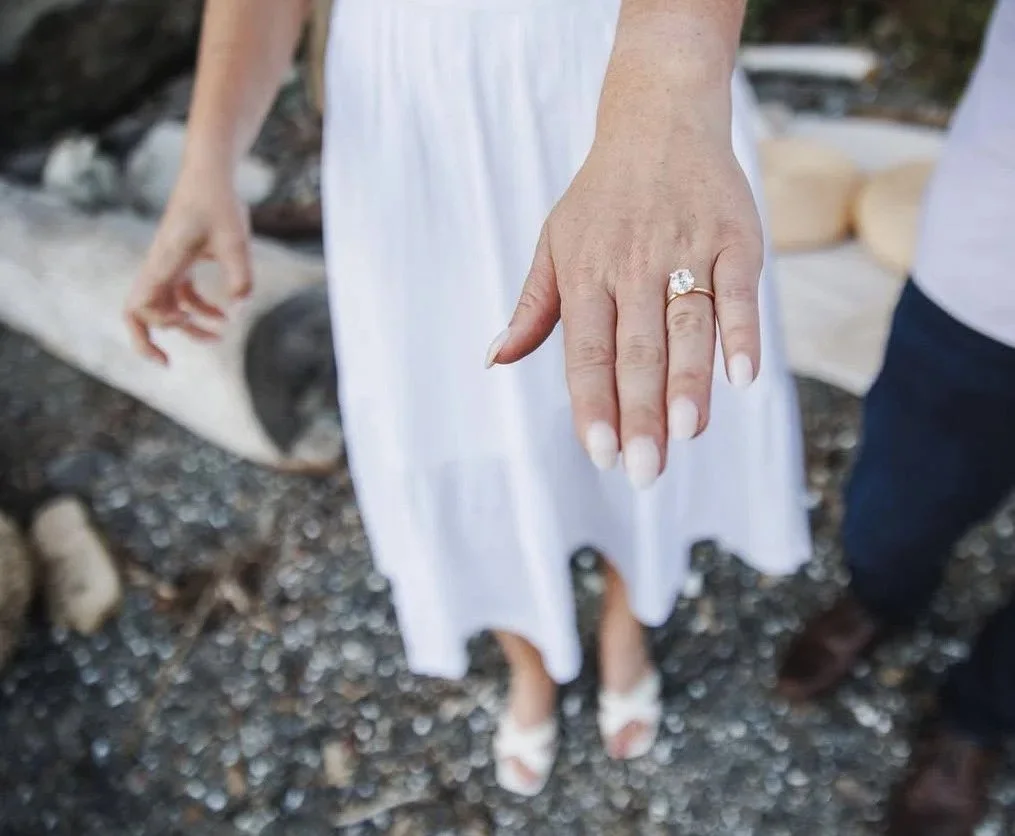 Close-up of a woman's left hand showing an engagement ring with a large diamond, with her nails painted white, on a woman wearing a white dress, standing outdoors with a rocky ground.