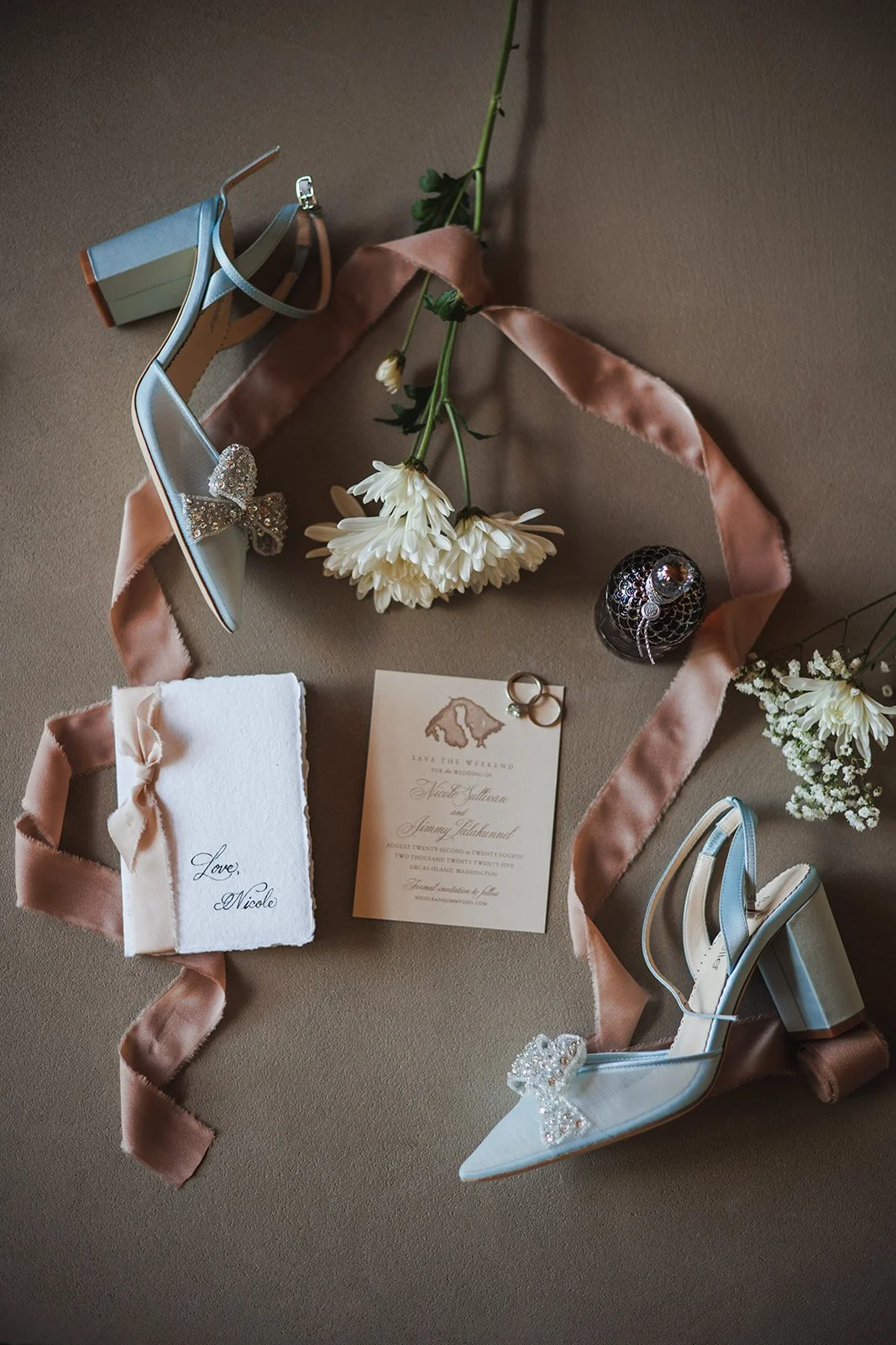 Wedding accessories including white high-heeled shoes with bow embellishments, a pink ribbon, white flowers, wedding rings, an invitation card, and a small gift box with a ribbon on a beige surface.