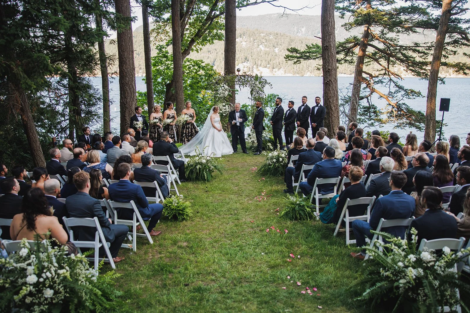 A wedding ceremony outdoors by a lake, with the bride and groom standing before a officiant. Bridesmaids and groomsmen stand beside them, and guests are seated in white chairs on either side, surrounded by trees and overlooking the water.