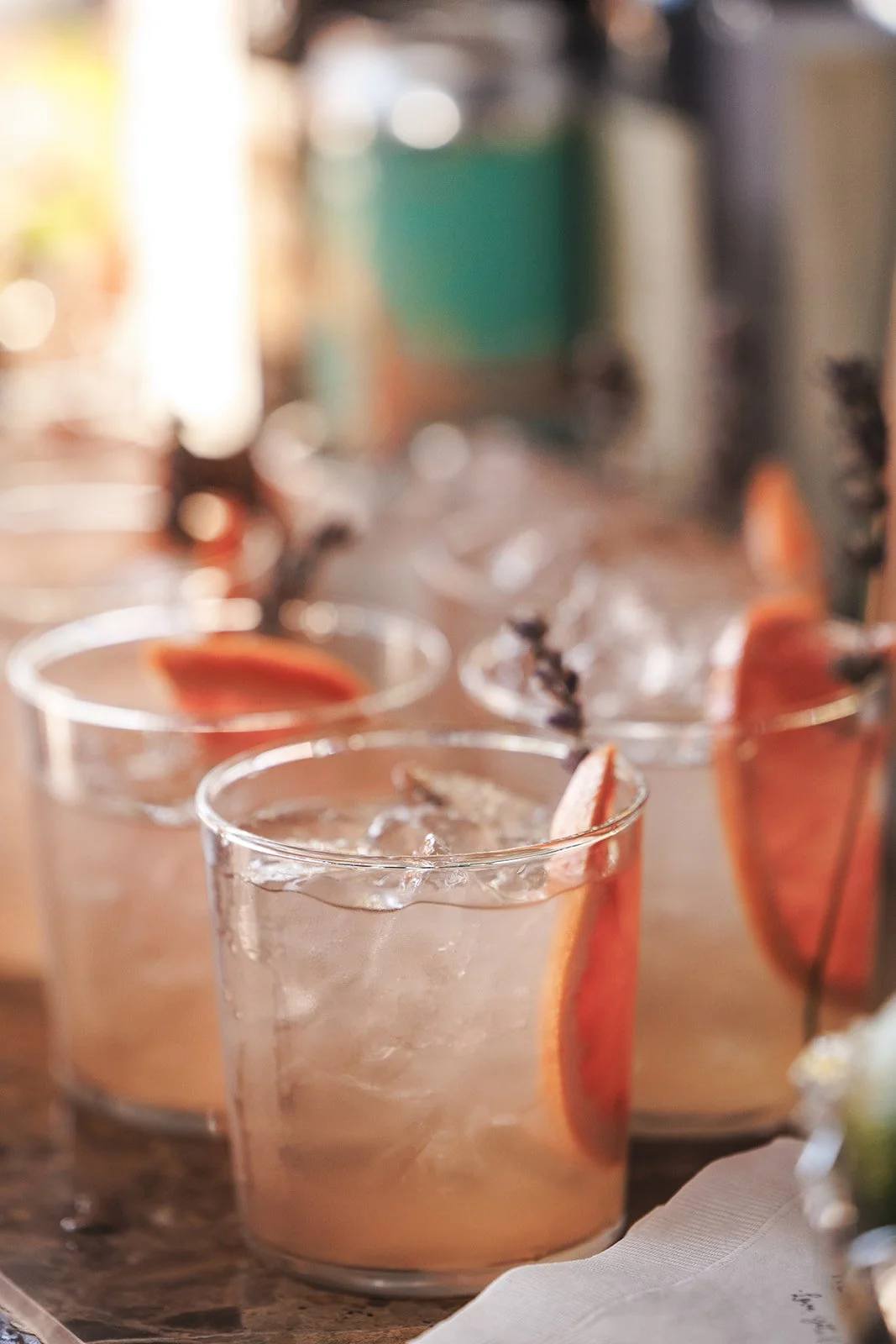 Three glasses of a pinkish cocktail with orange slices and sprigs of lavender, on a wooden surface