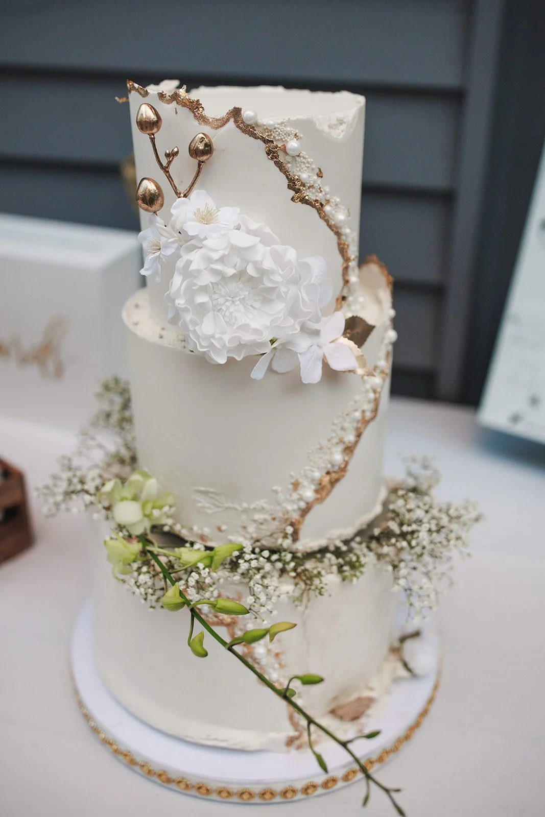 Two-tiered white wedding cake decorated with gold accents, white flowers, and greenery.