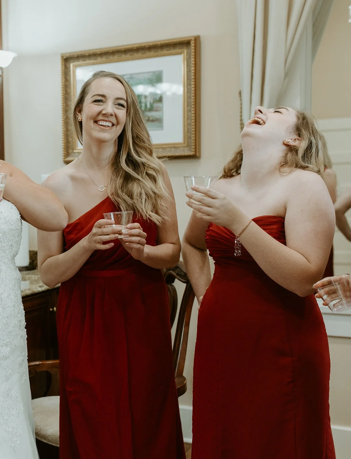 Two women in red strapless dresses smiling and laughing at an indoor gathering, holding clear plastic cups.