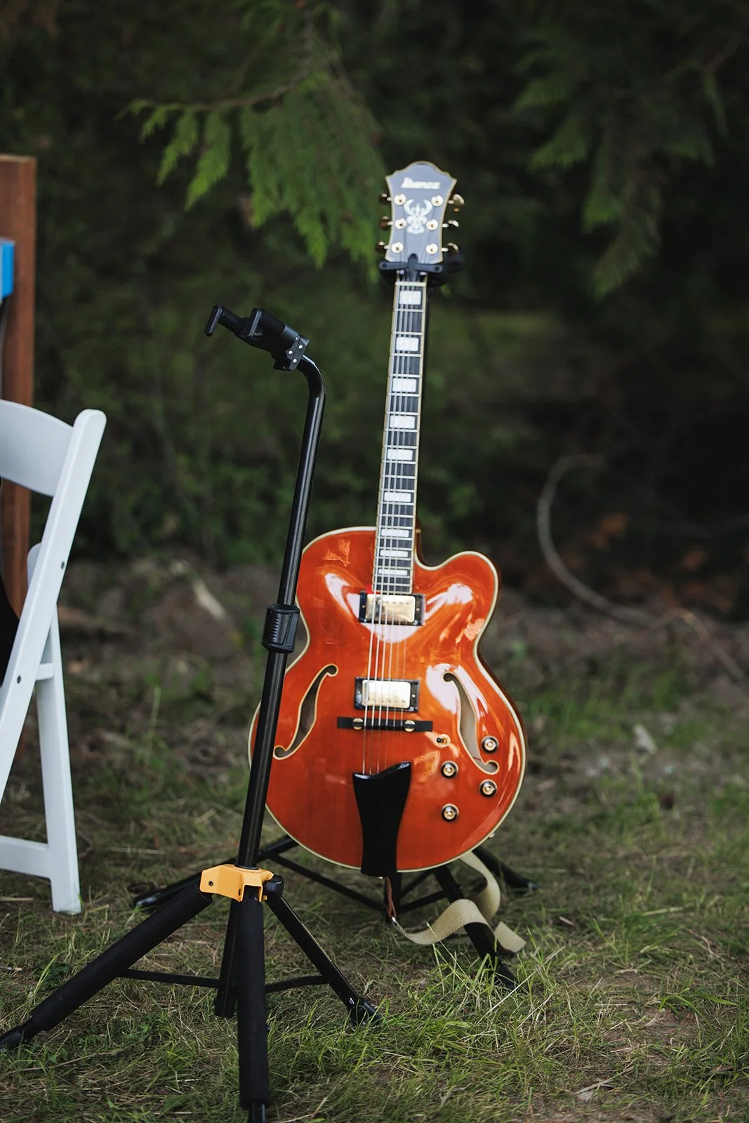 An orange hollow body electric guitar standing on a stand outdoors, with a white chair partly visible to the left and a forest background.