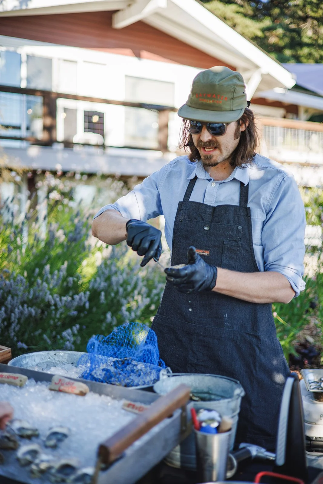 A man wearing sunglasses, a cap, a light blue shirt, a black apron, and black gloves prepares seafood at an outdoor seafood market or restaurant, with a house and greenery in the background.