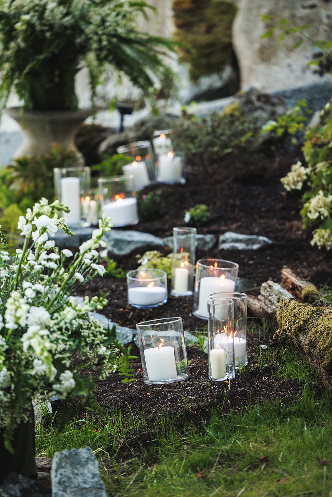  Candlelit outdoor memorial with white candles in glass holders and white flowers in a garden setting.