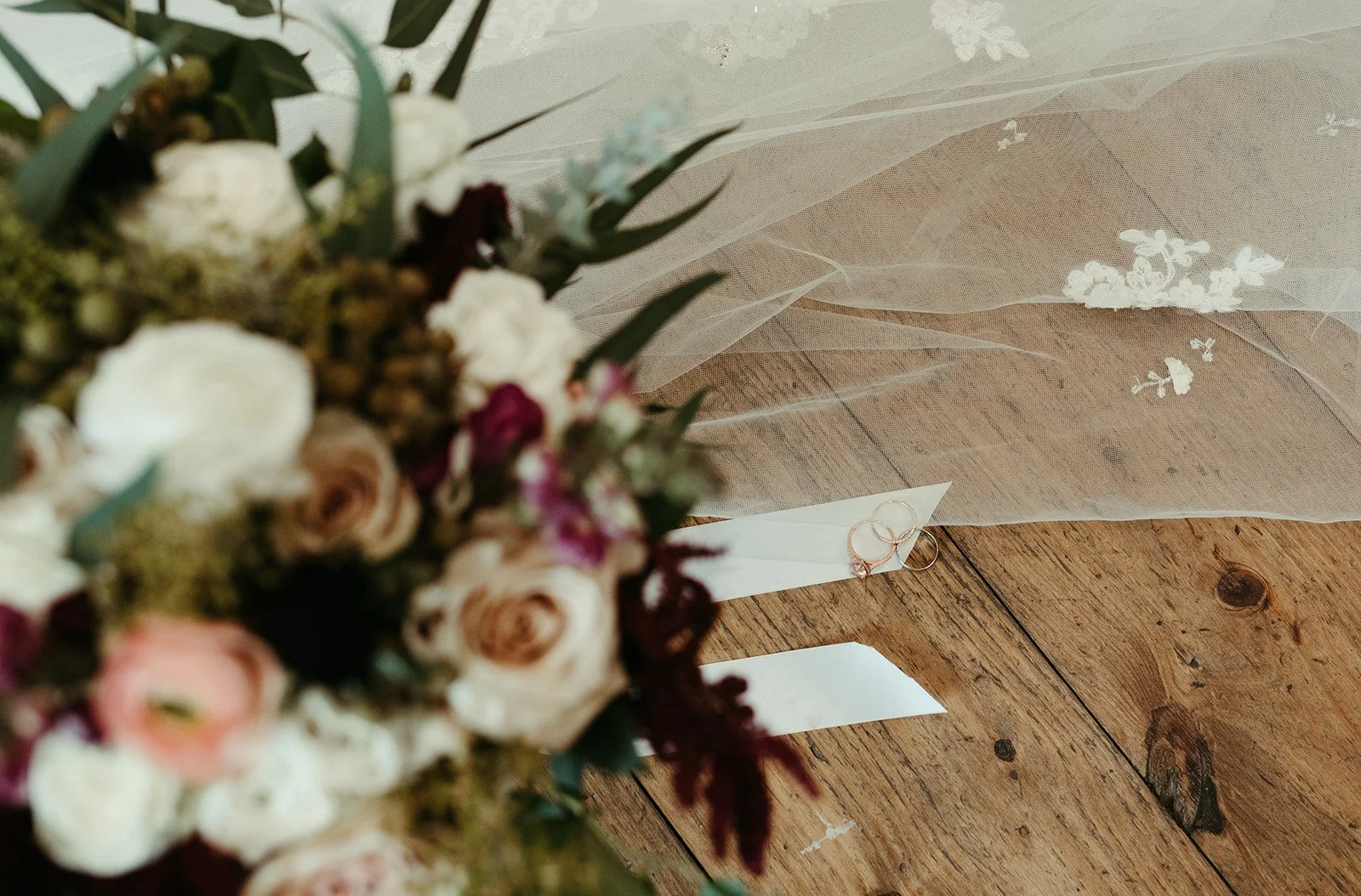 A wedding bouquet with white, pink, and burgundy flowers and greenery, resting on a wooden floor next to a sheer white veil with floral embroidery and two gold wedding rings.