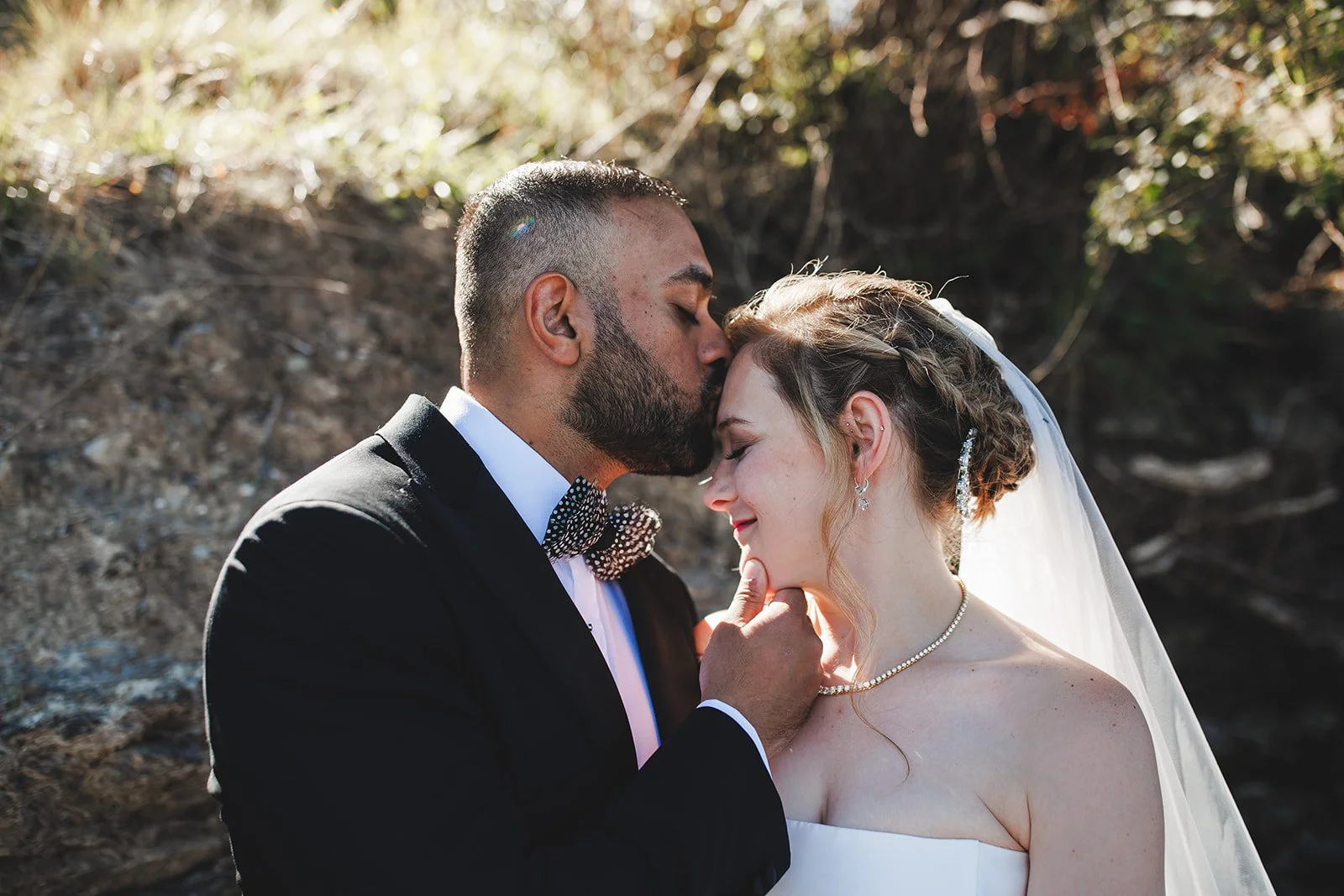 A groom kissing a bride on the forehead outdoors during a wedding, with a dirt and foliage background.