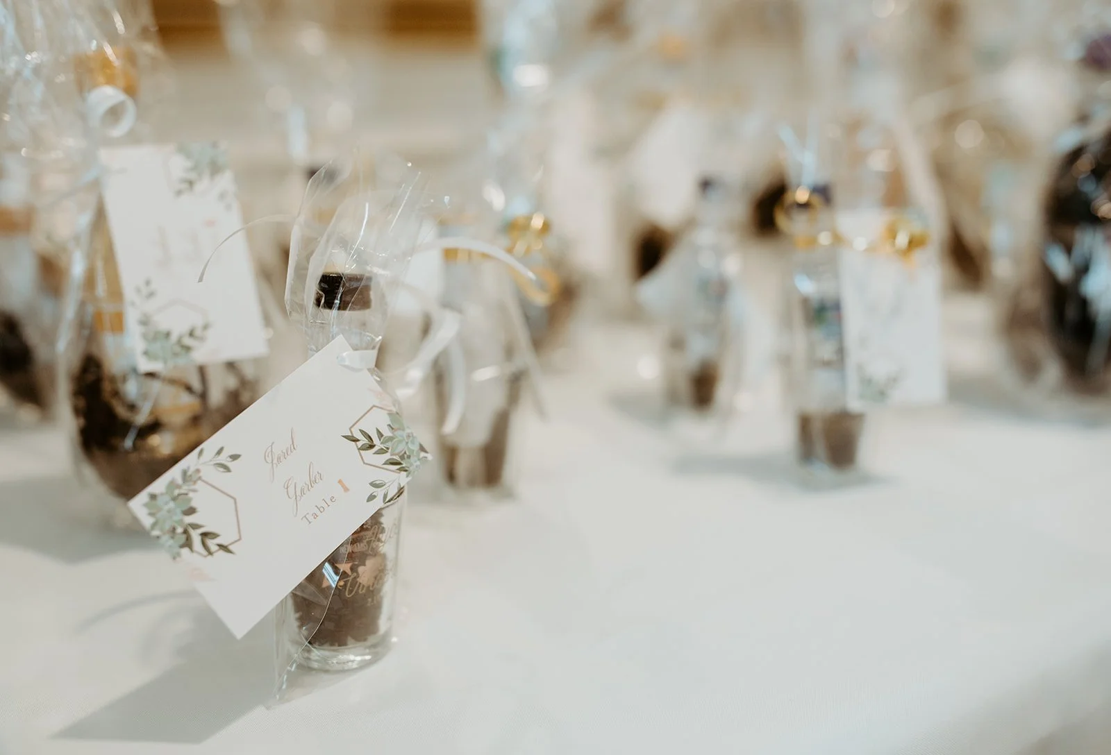Small glass jars wrapped in clear plastic with tags and floral designs, set on a white table, labeled for a wedding table named 'Garden Table 1'.