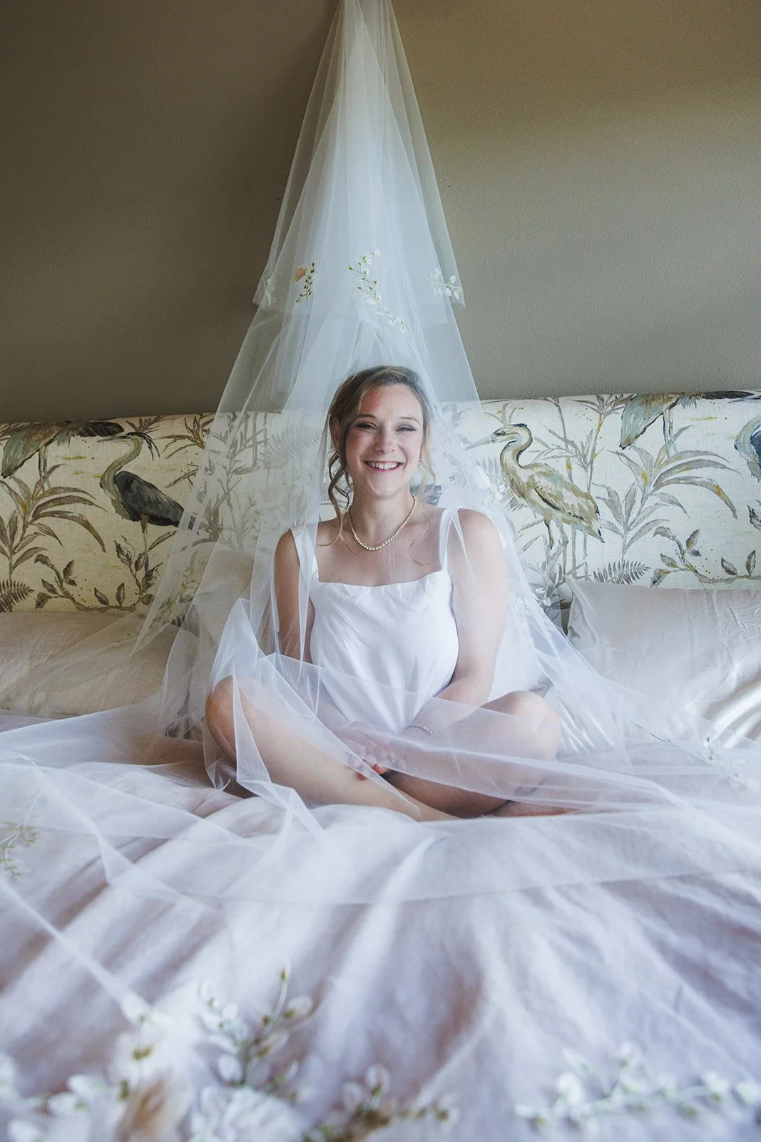 Smiling young woman sitting on a bed, surrounded by sheer white fabric, with a botanical print headboard and a plain wall behind.