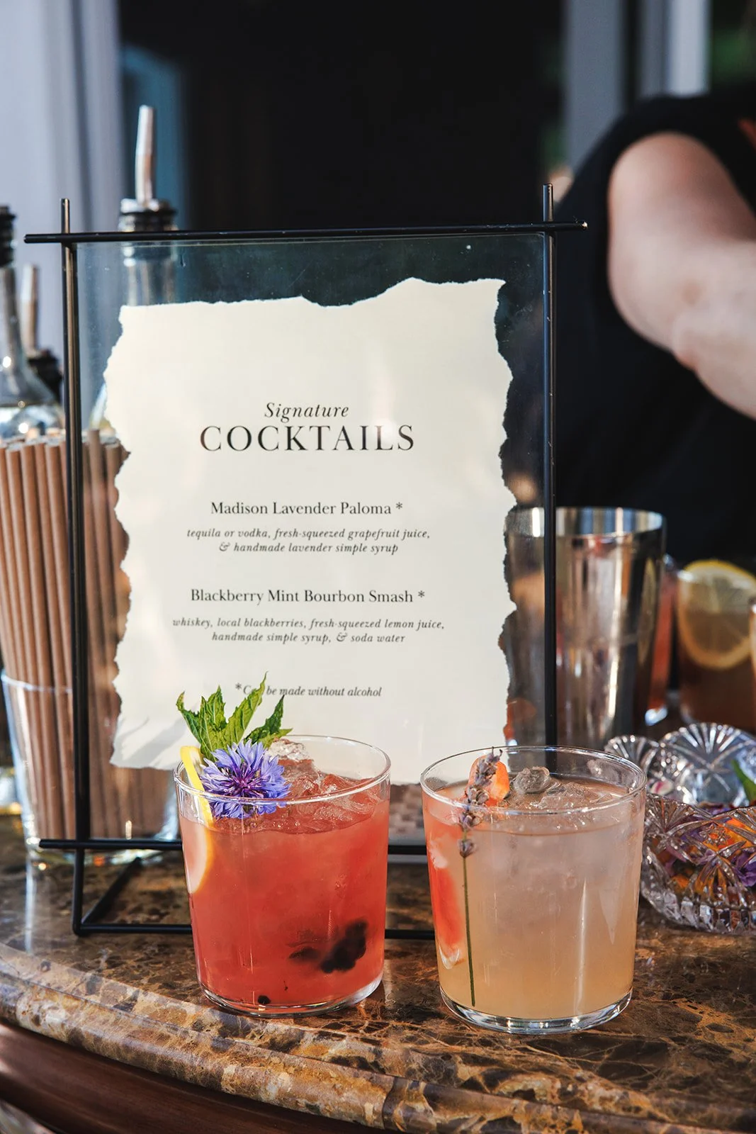 A menu sign for signature cocktails at a bar, with two colorful cocktails in front garnished with flowers and herbs, on a marbled counter.
