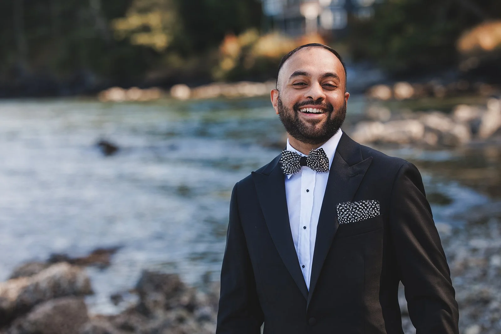 A man dressed in a tuxedo with a polka dot bow tie and pocket square, standing outdoors near a river with rocks and trees in the background, smiling.