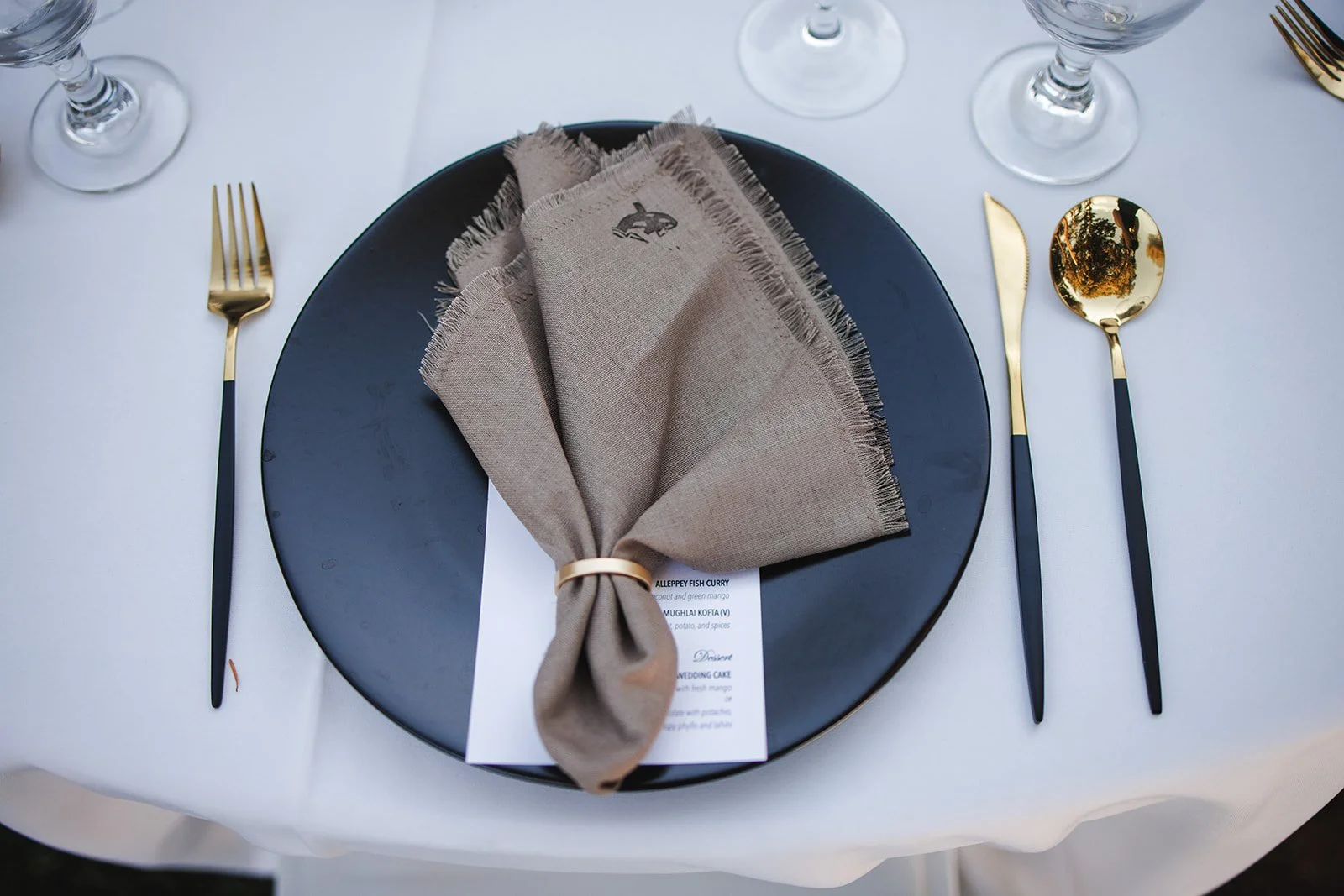 Elegant table setting with a black plate, gold and black flatware, a beige cloth napkin folded on the plate, and clear wine glasses on a white tablecloth.