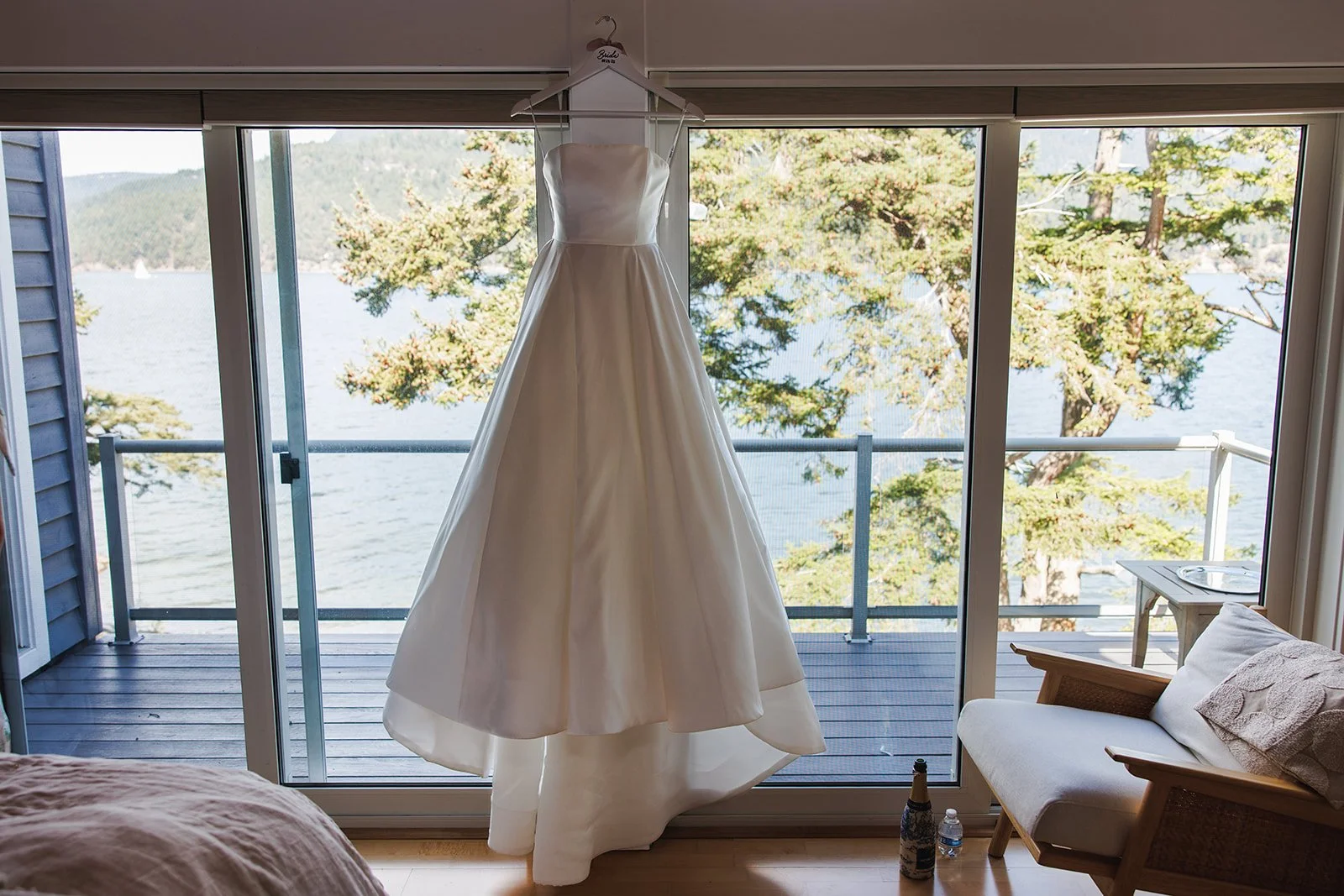 White wedding dress hanging on a hanger in front of large glass sliding doors overlooking a lake and trees.