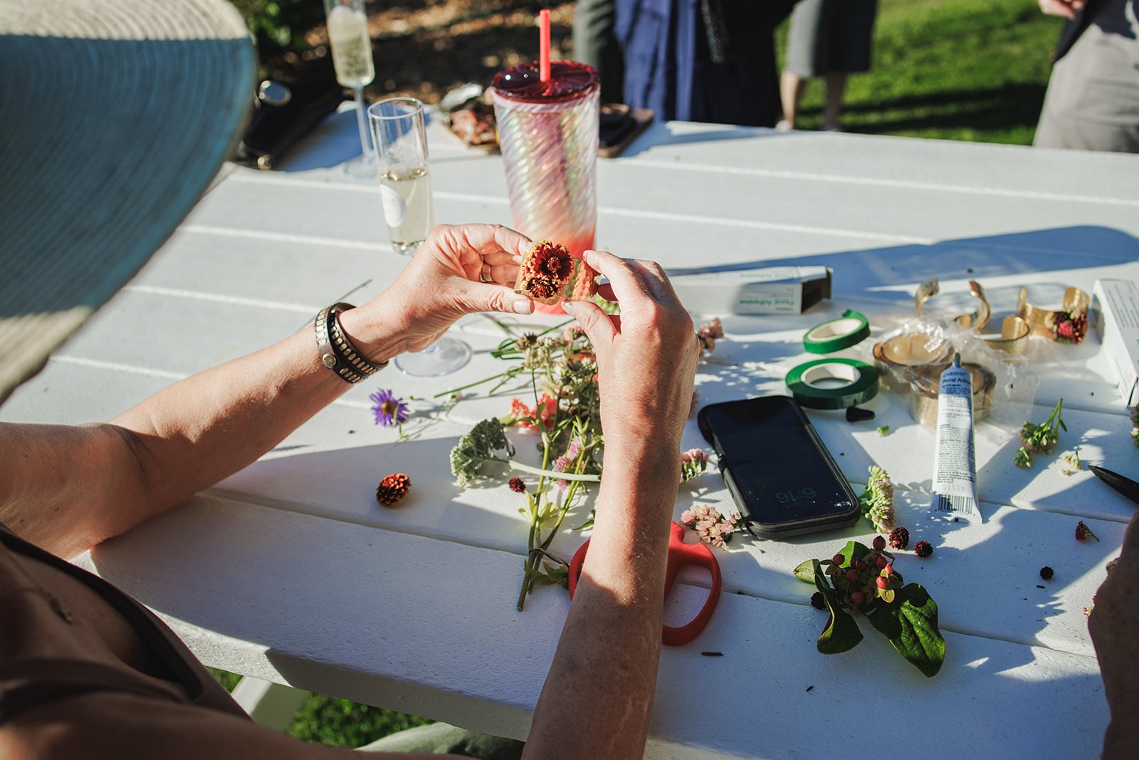 A person working on flower arrangements at an outdoor table with various flowers, tools, and craft supplies, during daylight.