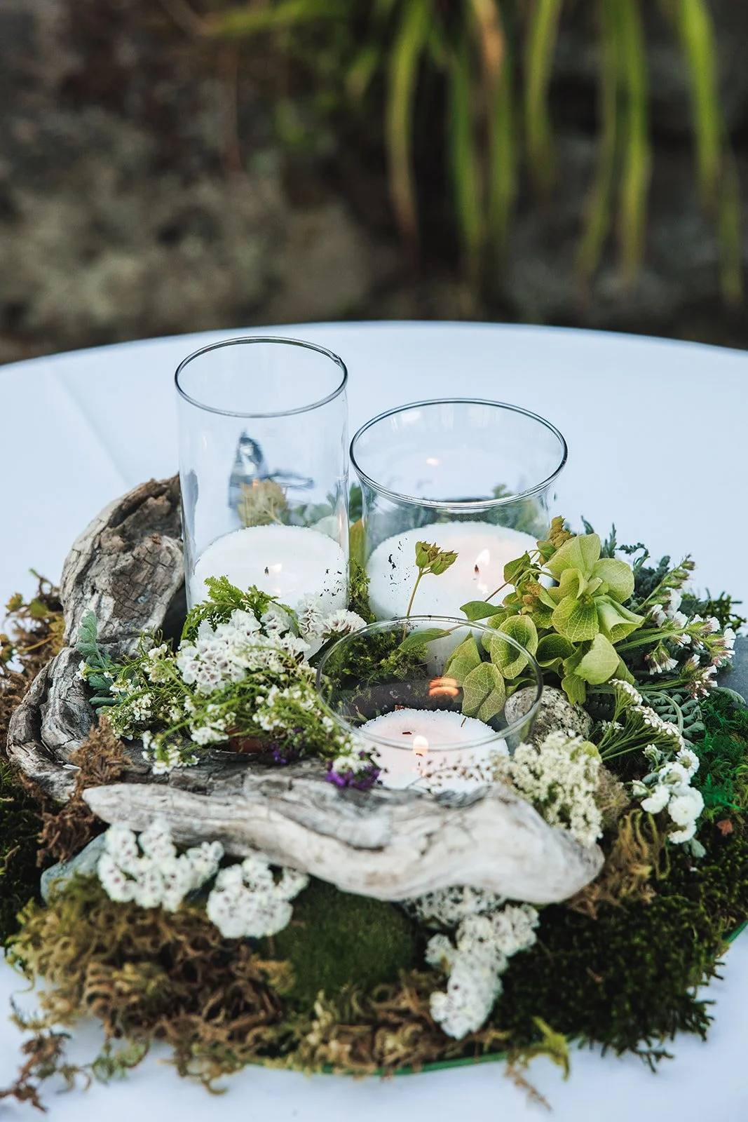 A decorative centerpiece with three glass candle holders containing candles, surrounded by green plants, flowers, moss, and driftwood on a white tablecloth.